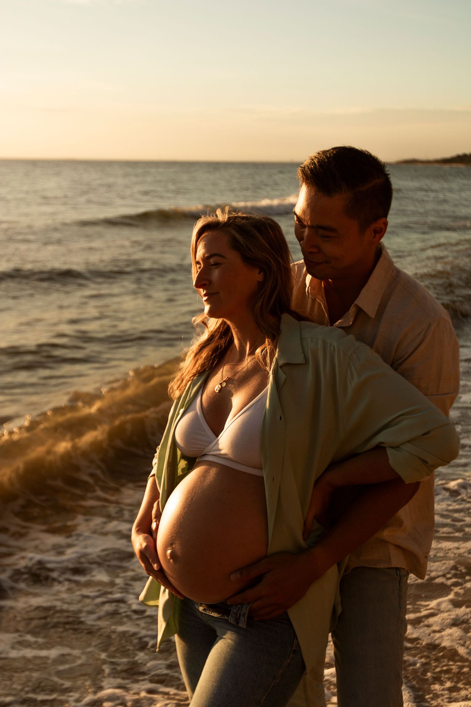 Romantic couple embraces on beach at sunset with gentle waves in background during golden hour maternity photography session.