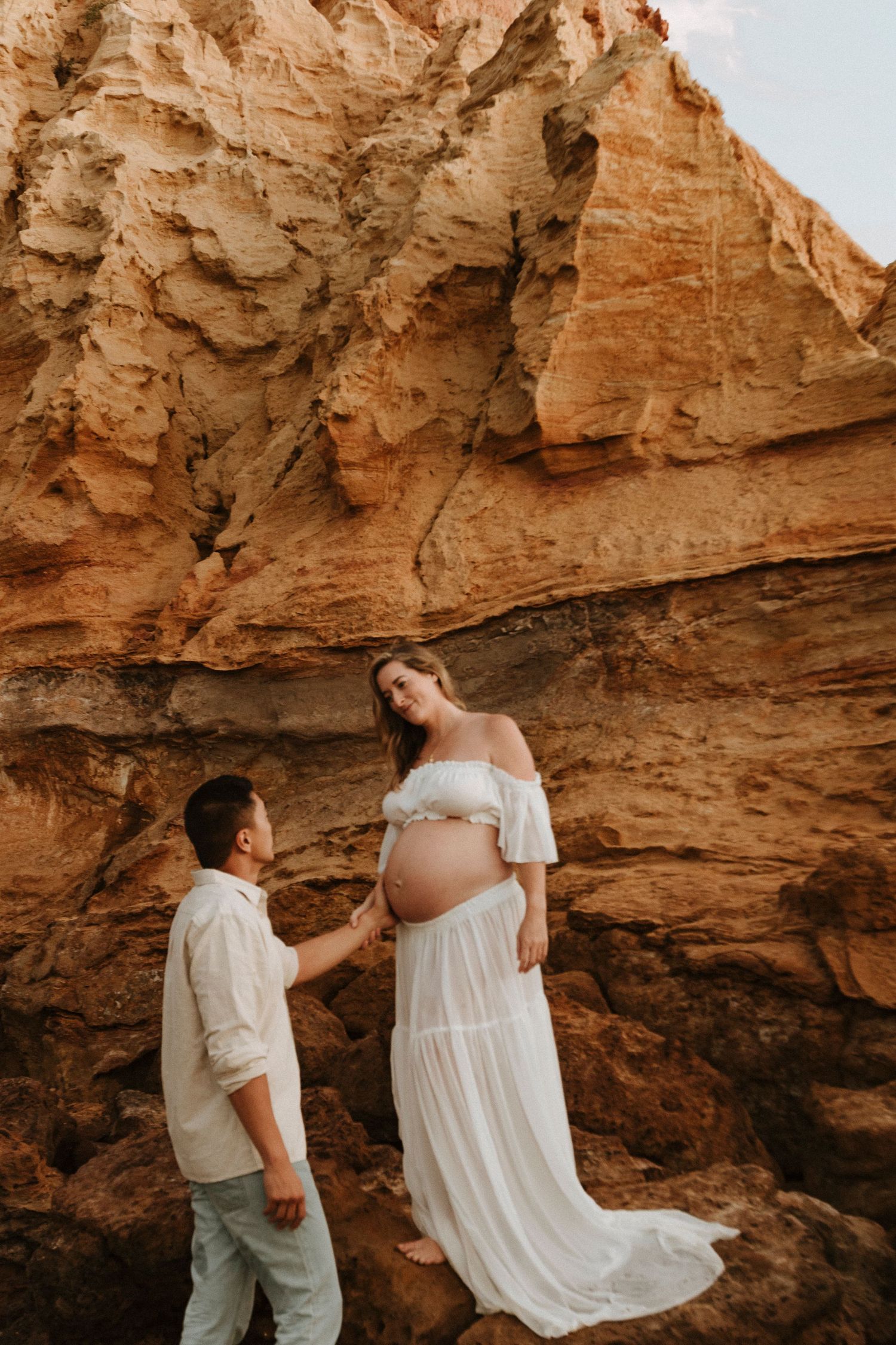 A romantic maternity photoshoot at sunset shows a couple posing against dramatic sandstone cliffs in golden light.