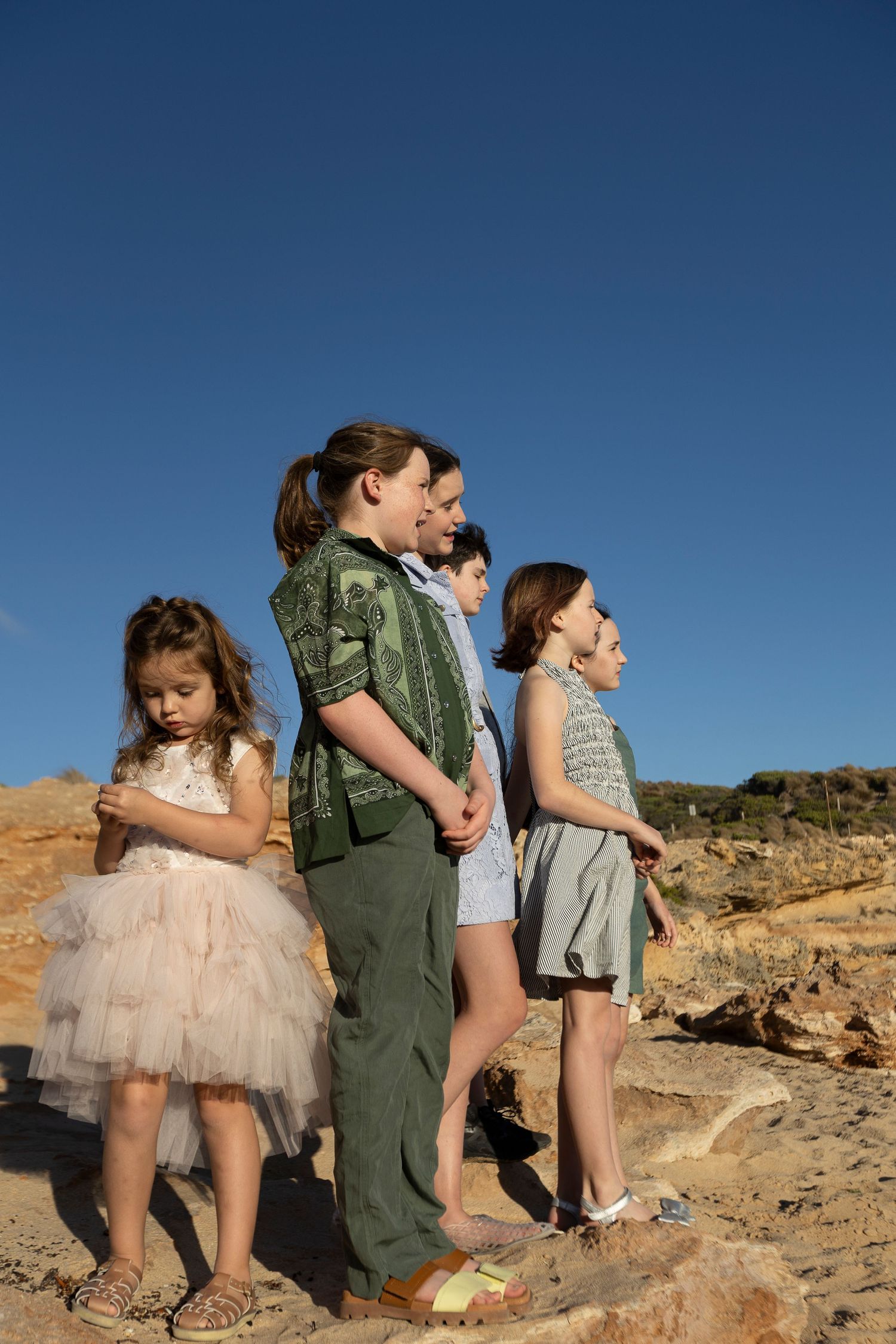 Family with children standing on rocky terrain under a clear blue sky, dressed in casual summer outfits.