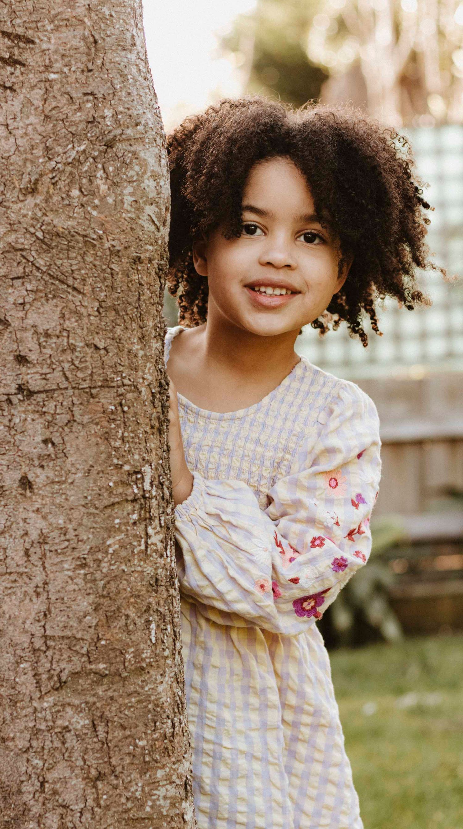 A child in a floral patterned dress peeks out from behind a tree trunk with natural curly hair and a bright smile.