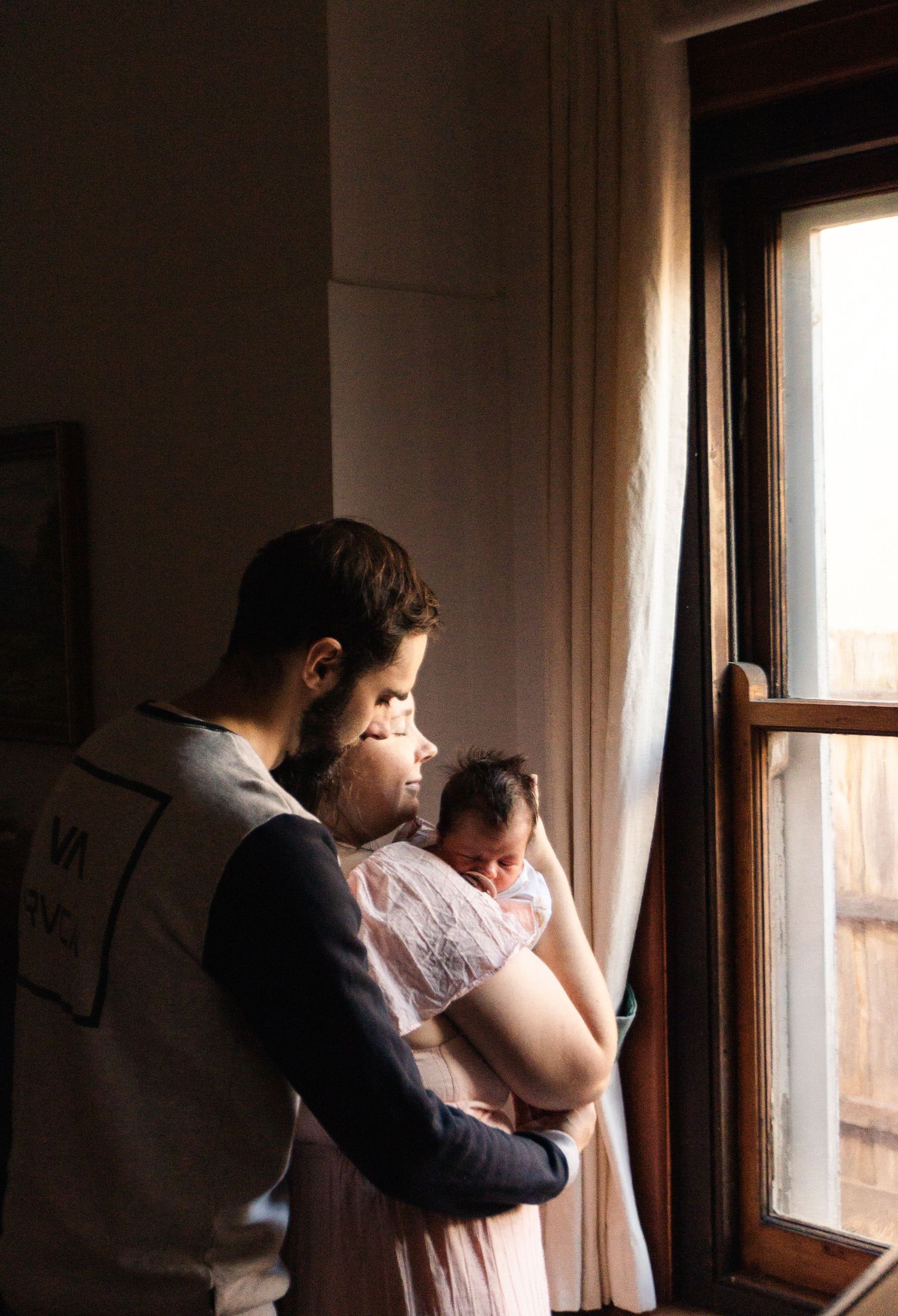 New parents standing by window in dimly lit room with natural light streaming in holding their newborn during a newborn photography session.