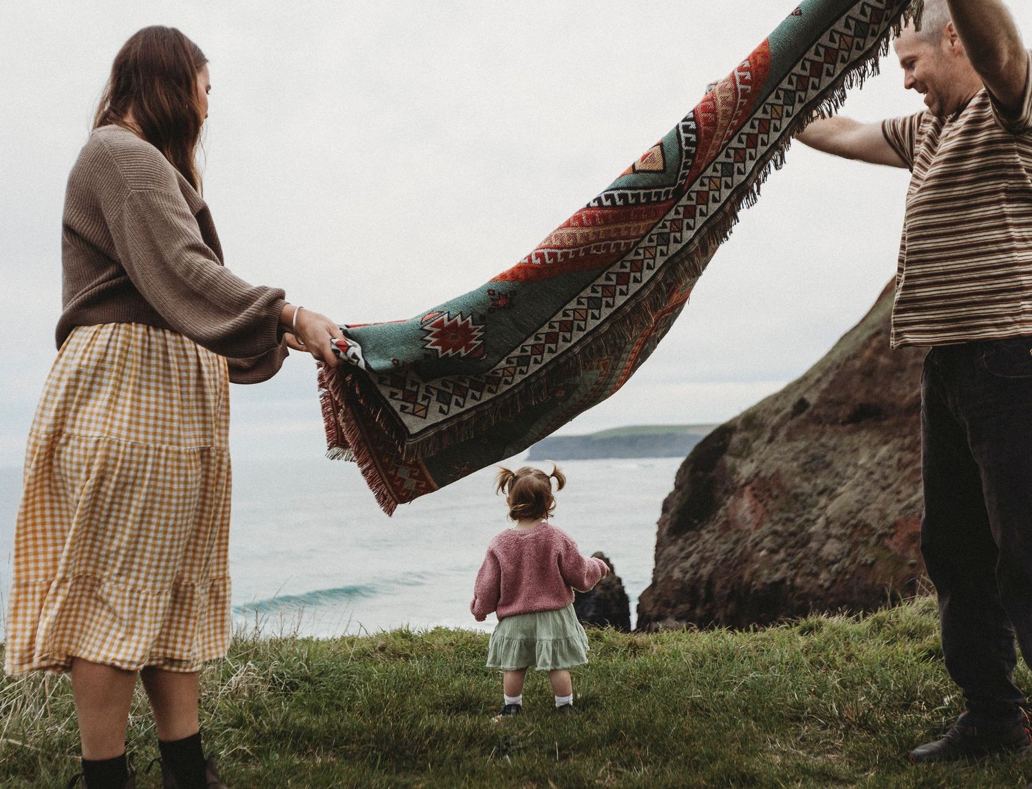 Family holding a colorful patterned blanket over a toddler near rocky coastal cliffs on a cloudy day.