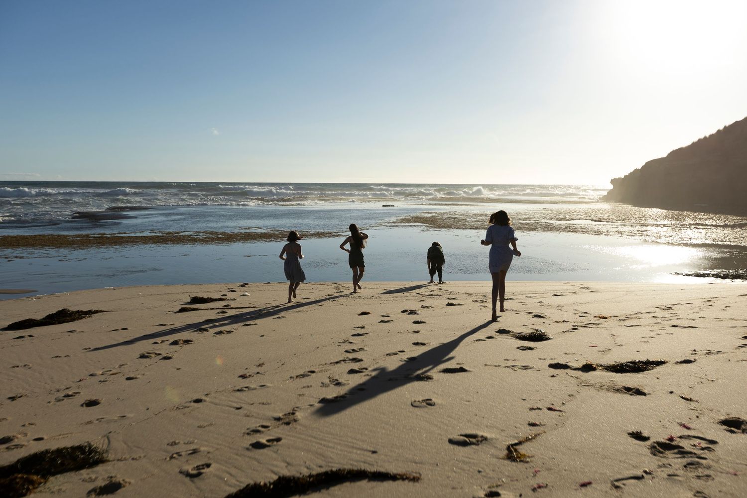 Four children running and playing on a sunny beach with ocean waves and golden sand in the background.