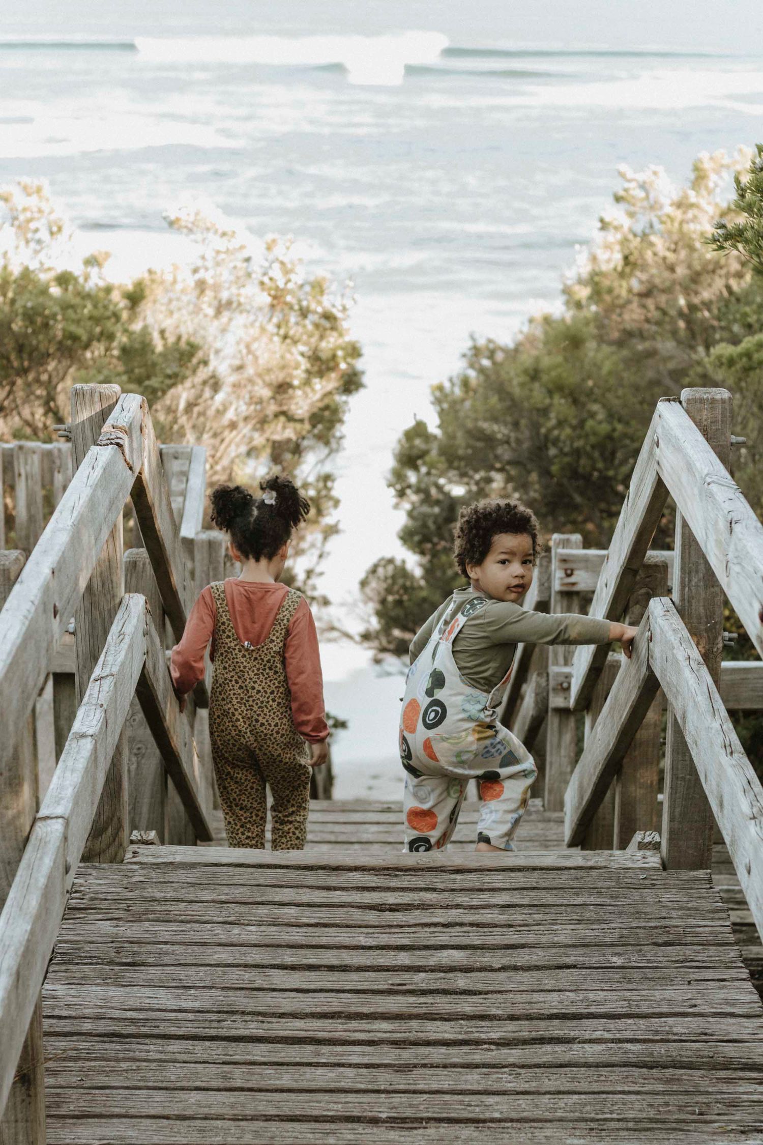 A beautiful family session shows two kids navigating down steep wooden stairs leading to a beach while surrounded by vegetation.