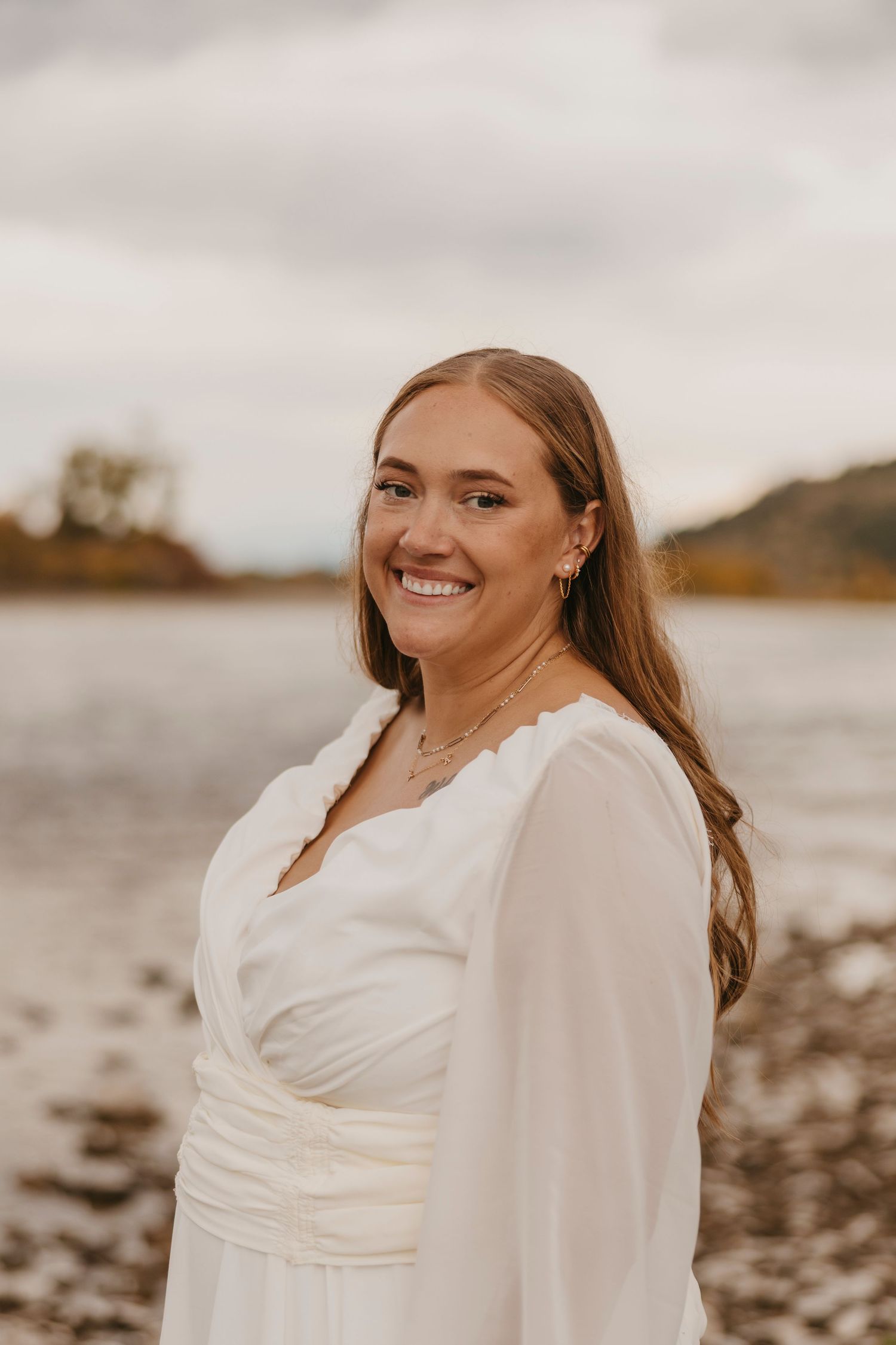 A person in a white dress stands by a lake shore at sunset with a warm smile and long hair flowing.