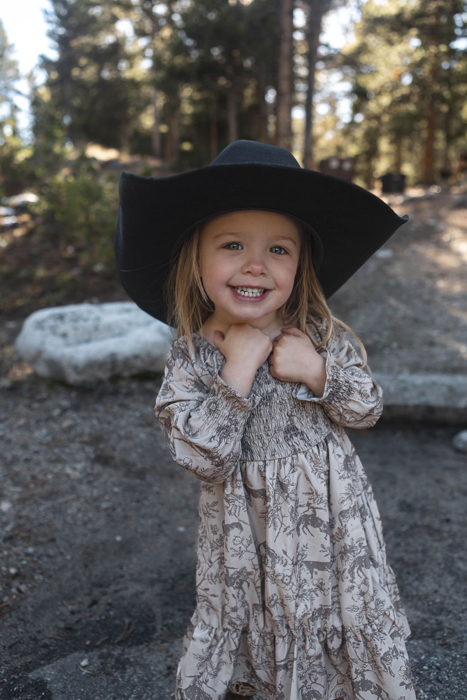 A child in a floral dress and black cowboy hat stands in a wooded outdoor setting with rocks and trees in the background.