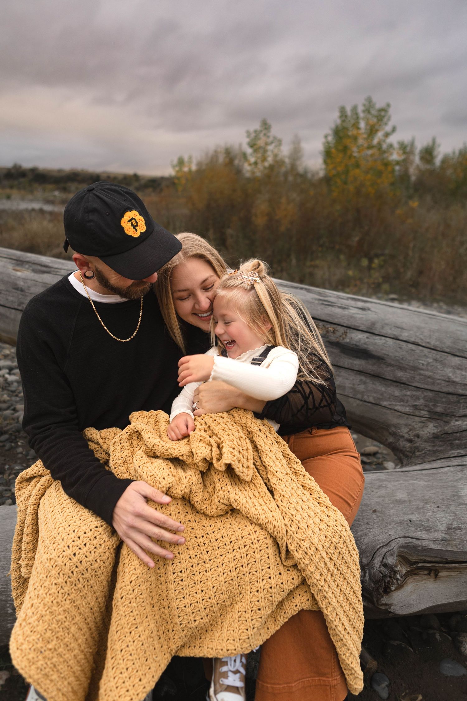 Couple wrapped in yellow knit blanket sharing intimate moments on driftwood at cloudy beach.