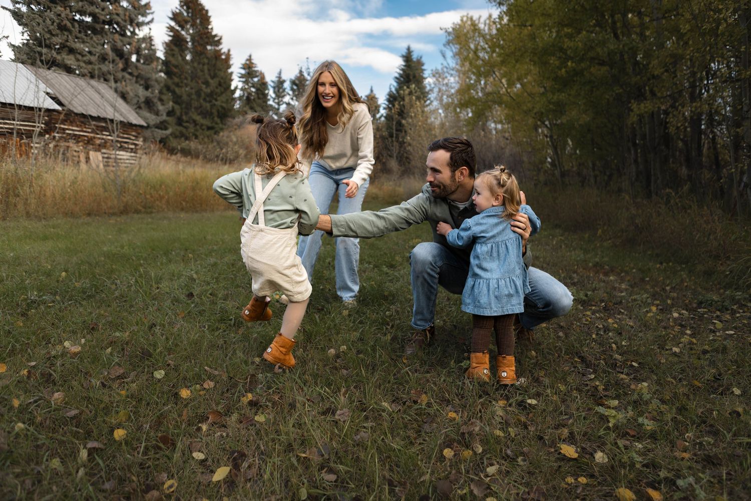 A family plays together outdoors in autumn, laughing and dancing in front of evergreen trees and a rustic building.