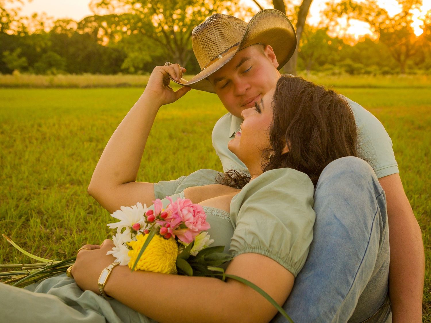 A couple relaxes in a grassy field at sunset with flowers beside them as golden light bathes the scene.