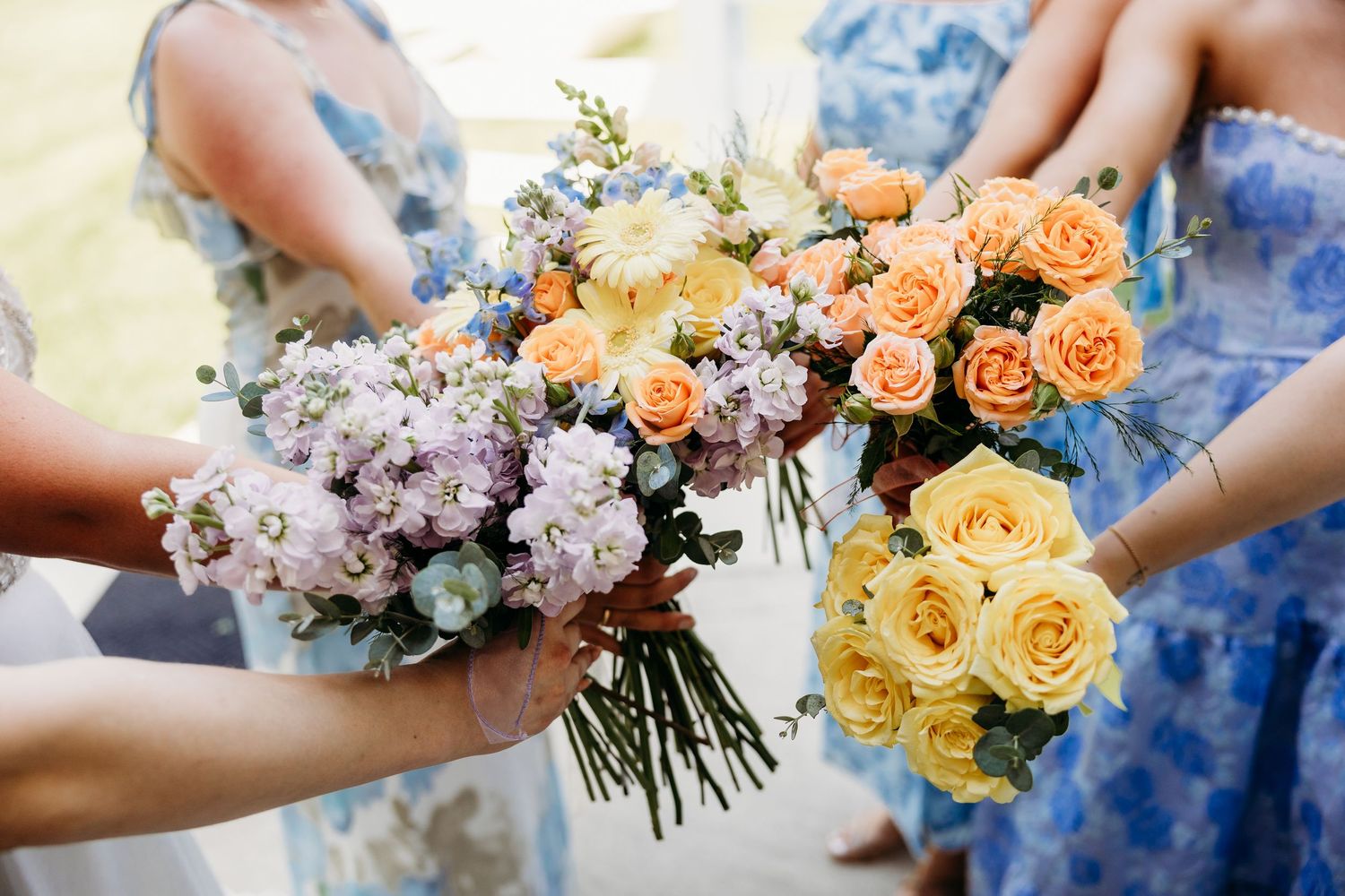 Bridesmaids in blue floral dresses holding bouquets of peach roses, yellow roses, and purple flowers at a wedding.