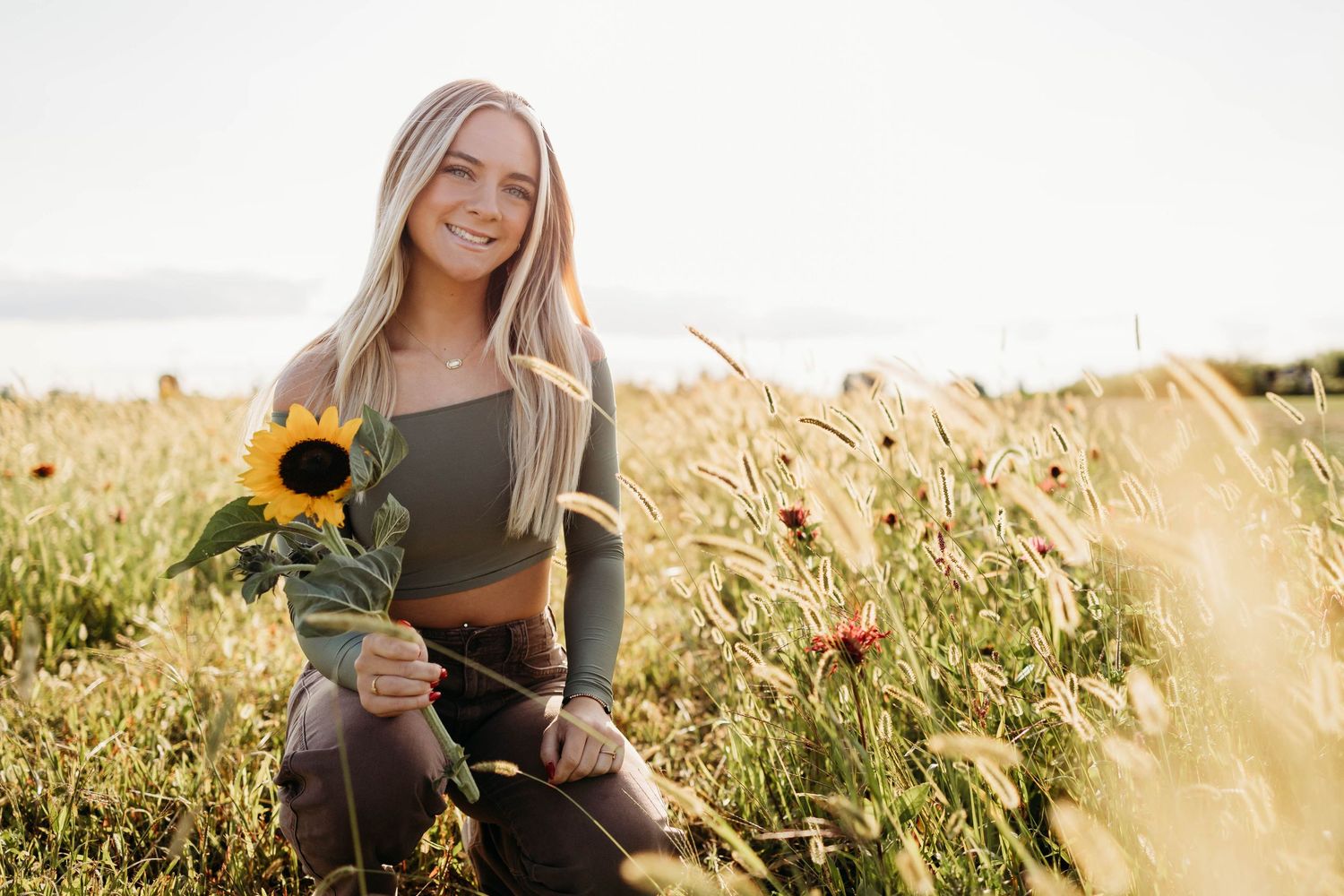 A person in a crop top sitting in a golden field at sunset holding a bright yellow sunflower.