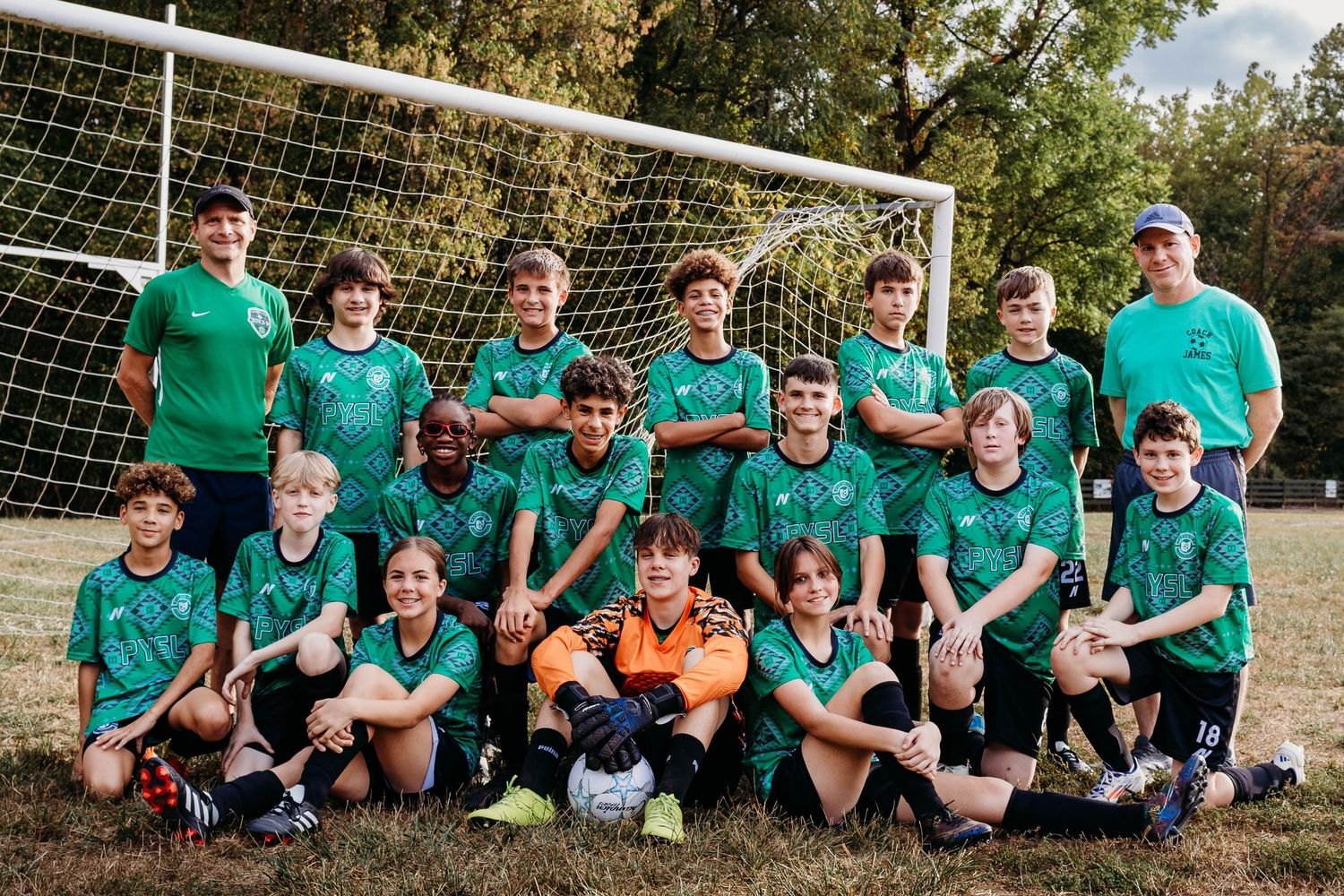 Youth soccer team in green jerseys poses together on field near goal while sitting on grass during sunny day.