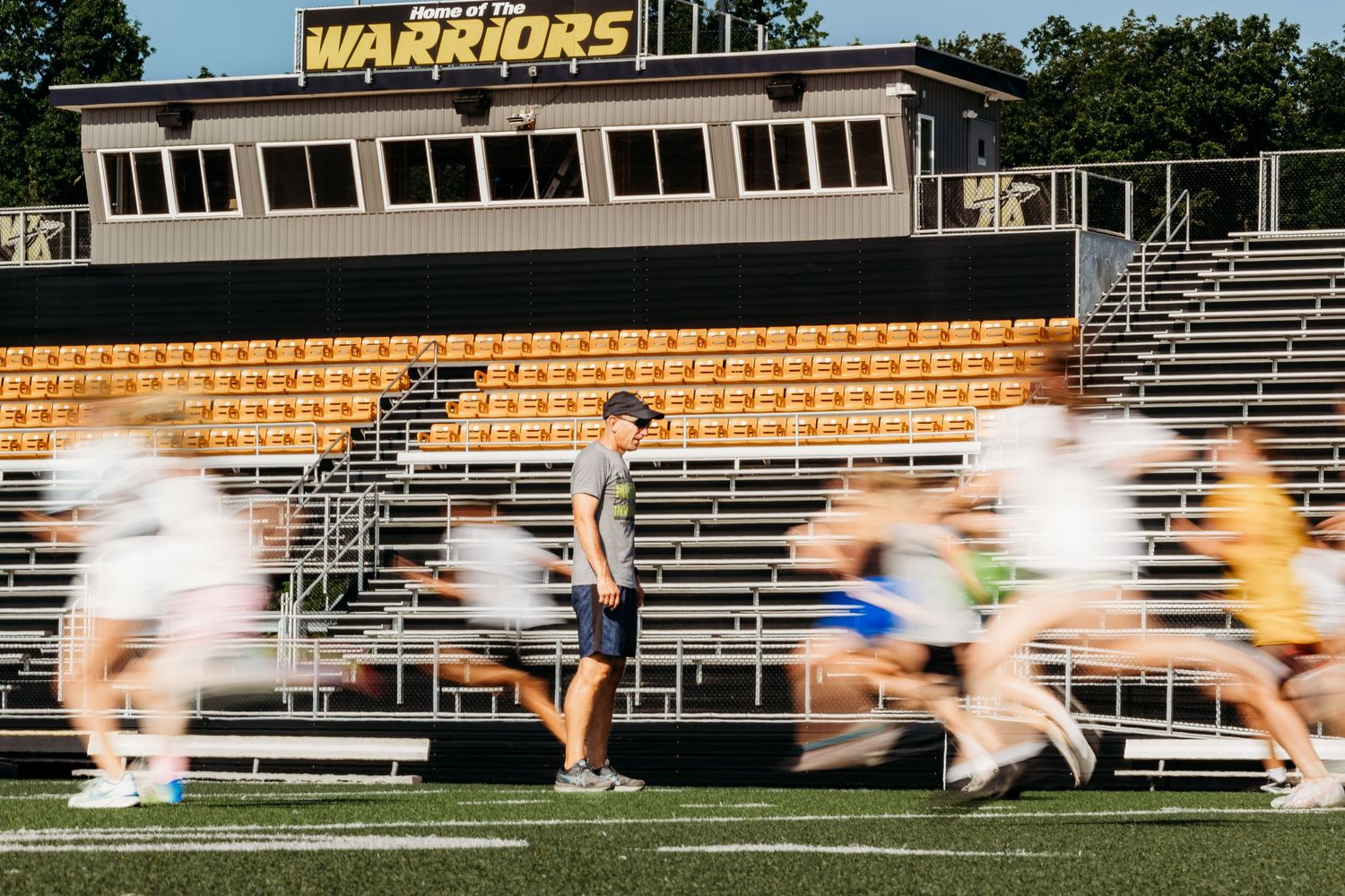 Motion blur captures a runner sprinting across a track field with orange stadium seats in the background.