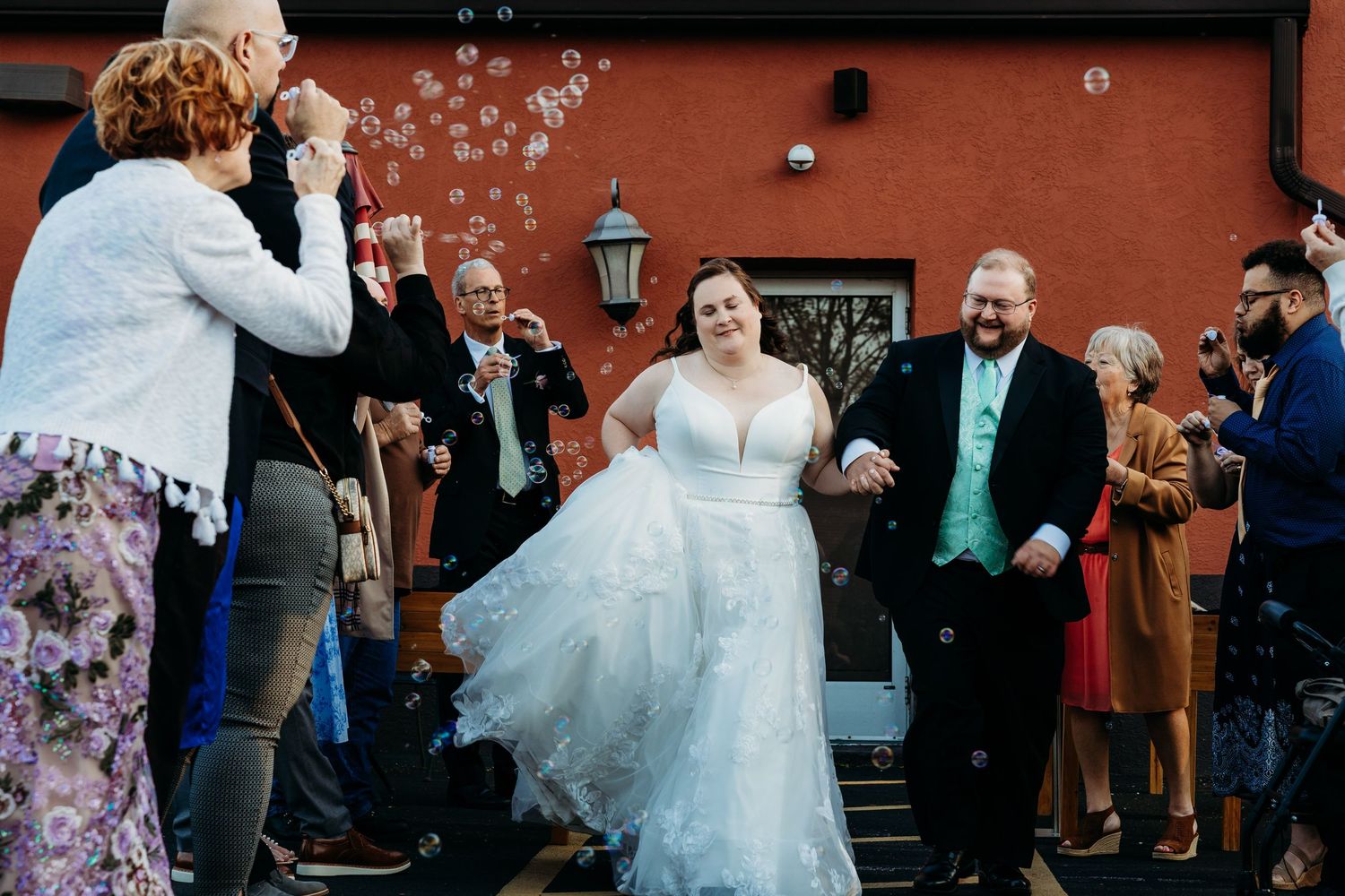 Wedding guests blow bubbles as couple exits down steps in joyful celebration at venue with red walls.
