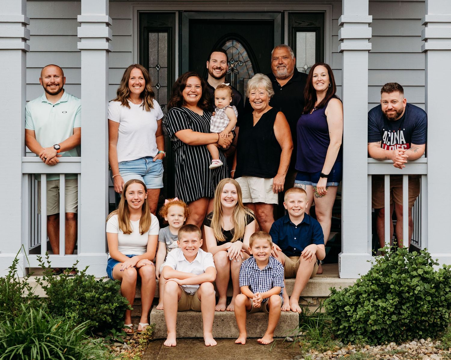 A large family group sits together on the steps of a white porch for a casual outdoor portrait.