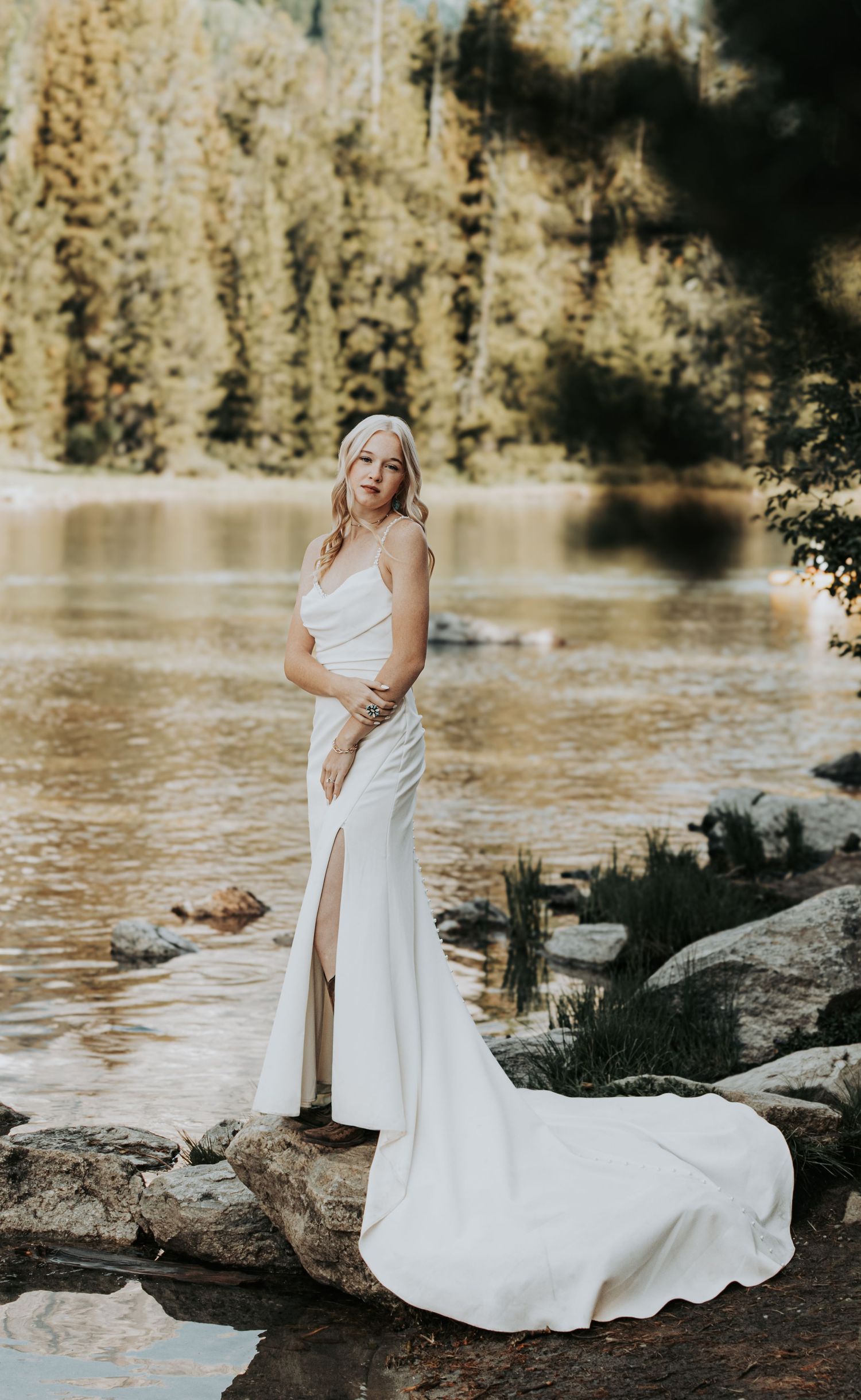A bride in a sleek white wedding dress with a thigh-high slit poses on rocks by a serene lake during golden hour.