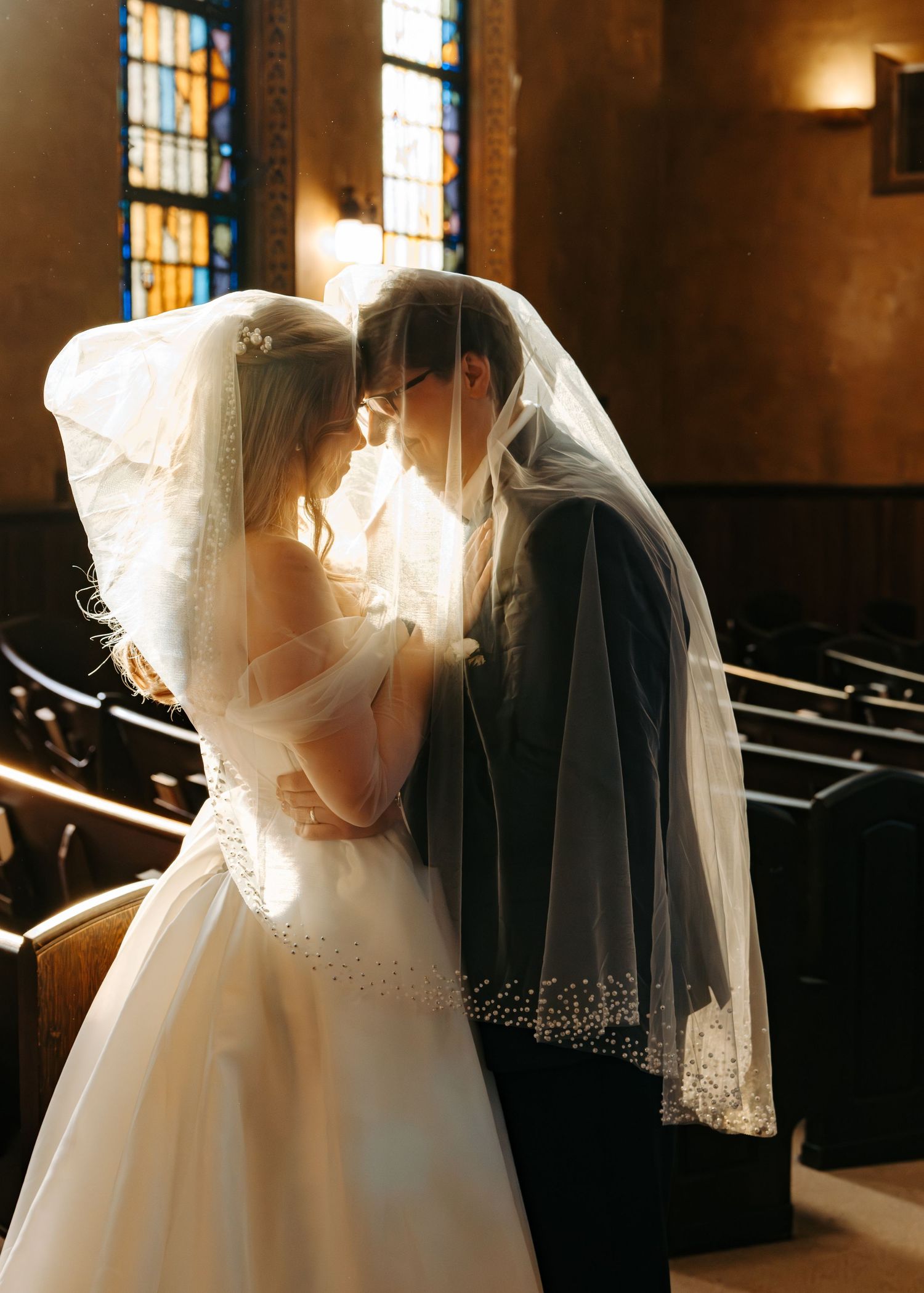 A romantic wedding scene in a church with sunlight streaming through stained glass windows, illuminating a tender moment.