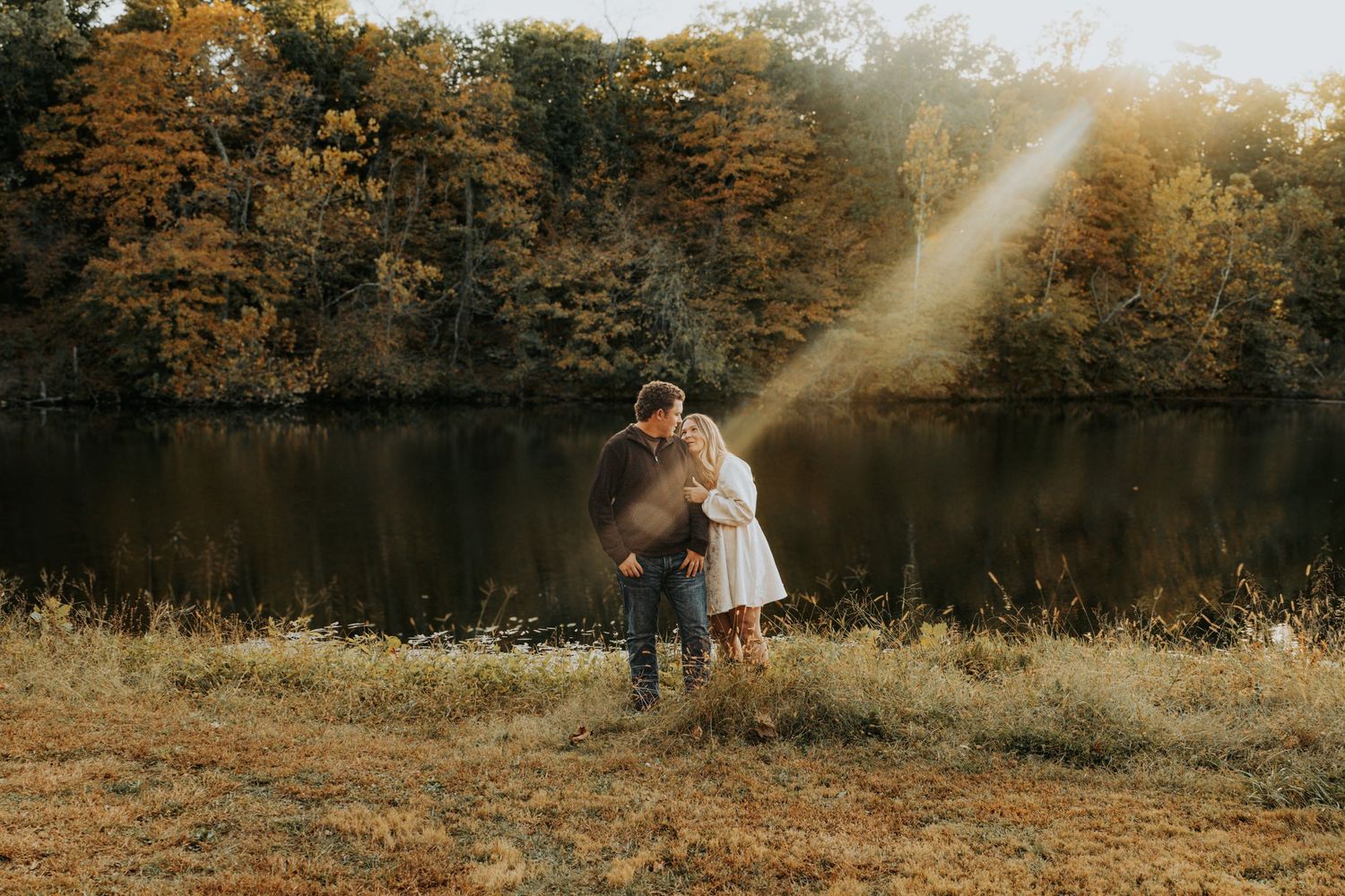 A couple embraces by a serene lake during golden hour as sunbeams shine through autumn trees.
