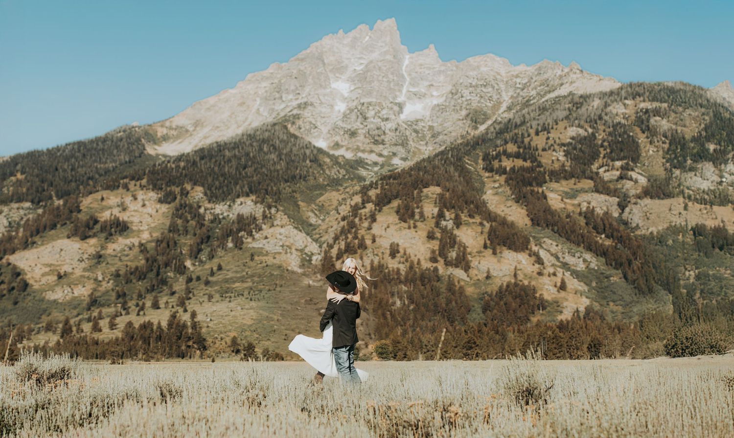 Panoramic mountain landscape sequence showing couple walking through golden meadow with dramatic peaks in background.