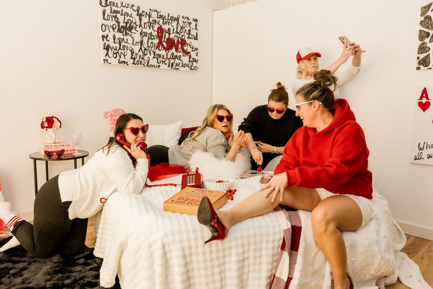 Four women celebrate Valentine's Day on a bed with red and white decor, gifts, and festive outfits.