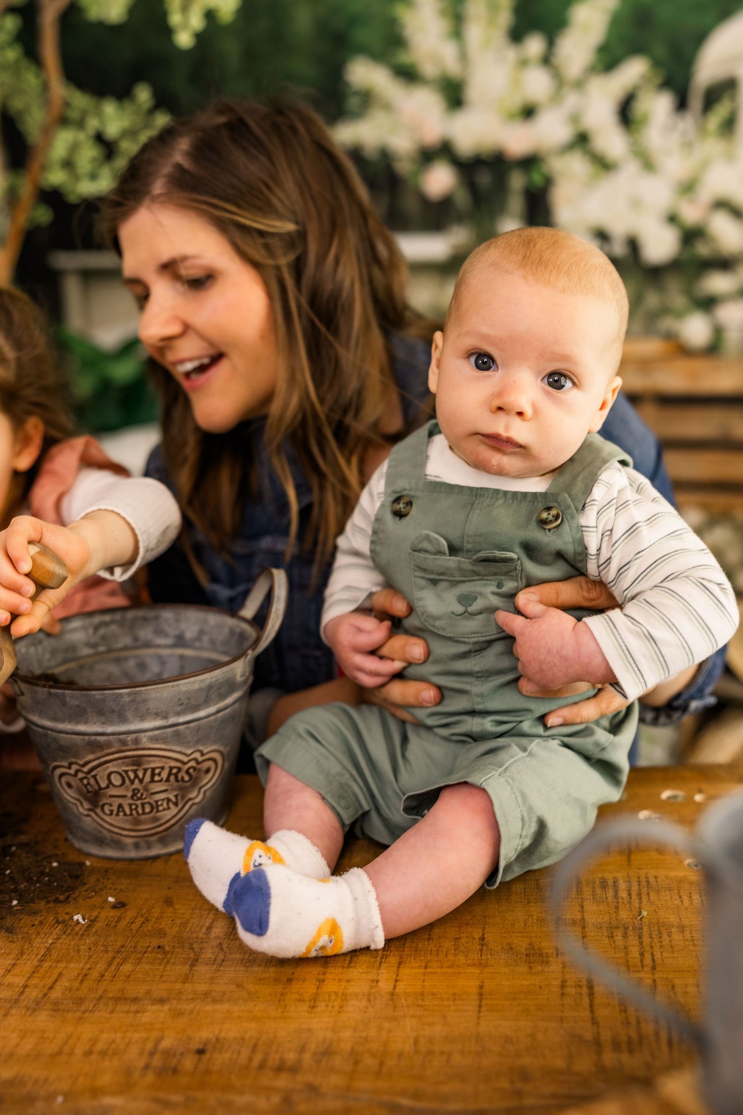 Baby in green overalls sits with a woman near a flower garden bucket in a cozy indoor setting.