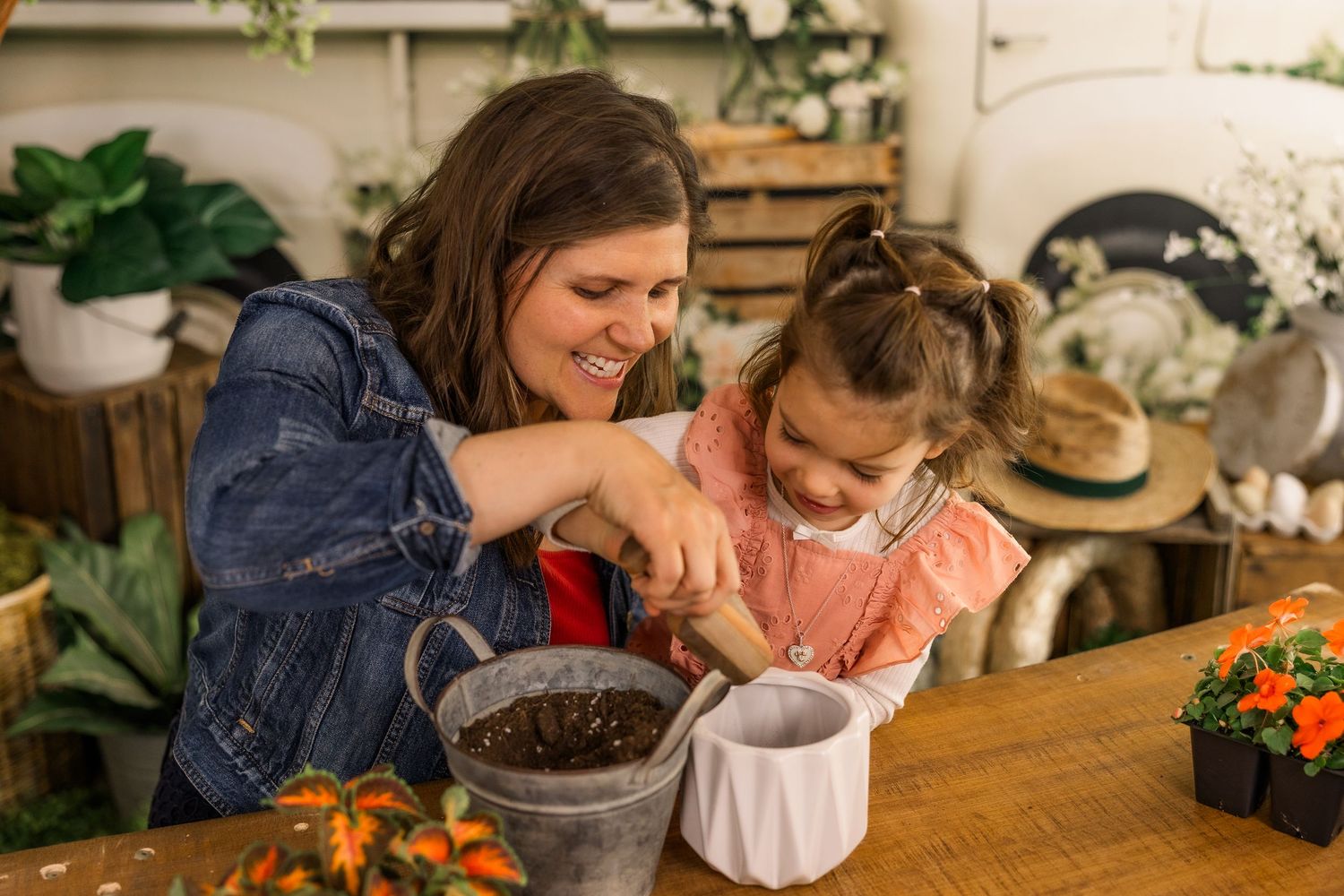 A mother and young daughter smile while potting soil into a white container together at a wooden table.