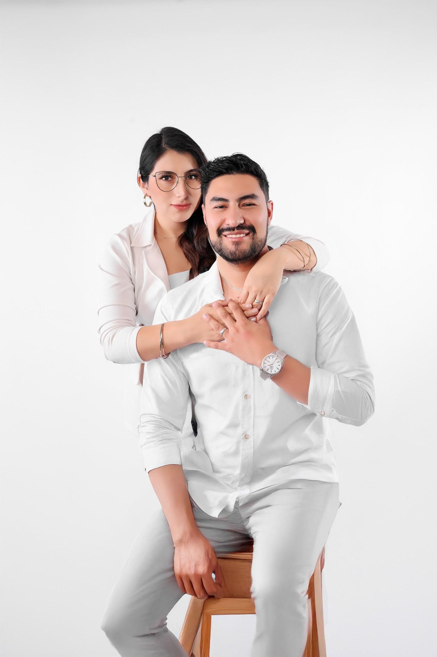 Smiling couple in white outfits posing together, woman with glasses hugging man seated on wooden chair.