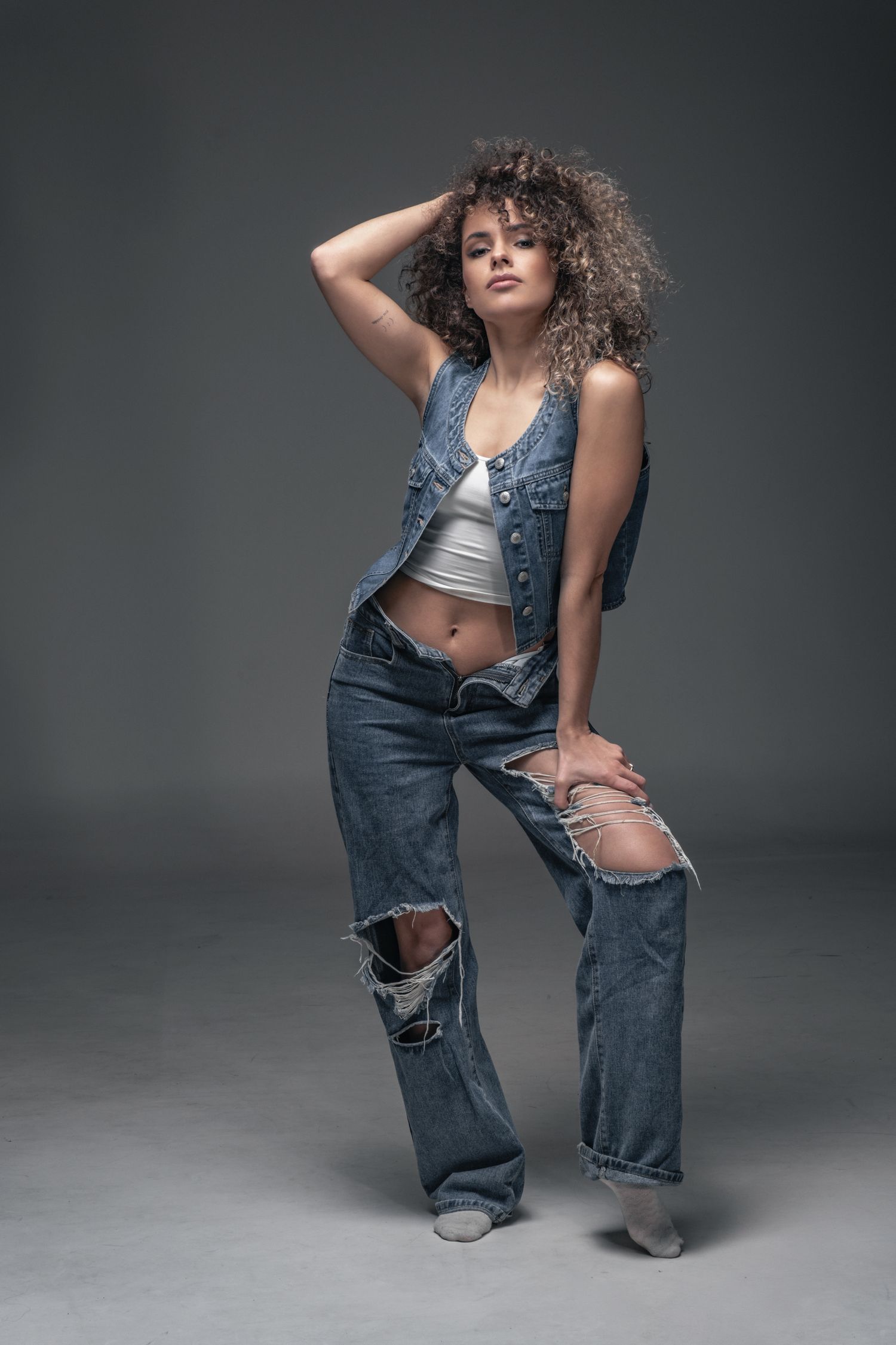 Full-body fashion editorial: model in distressed denim and vest, minimalist studio setting, high-contrast lighting.