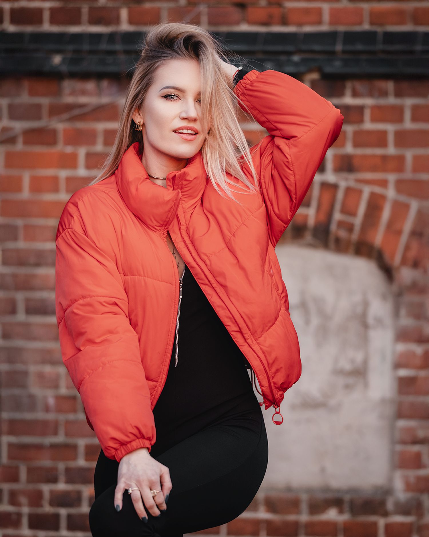 Blonde woman in red puffer jacket posing confidently against a red brick wall wearing black pants.