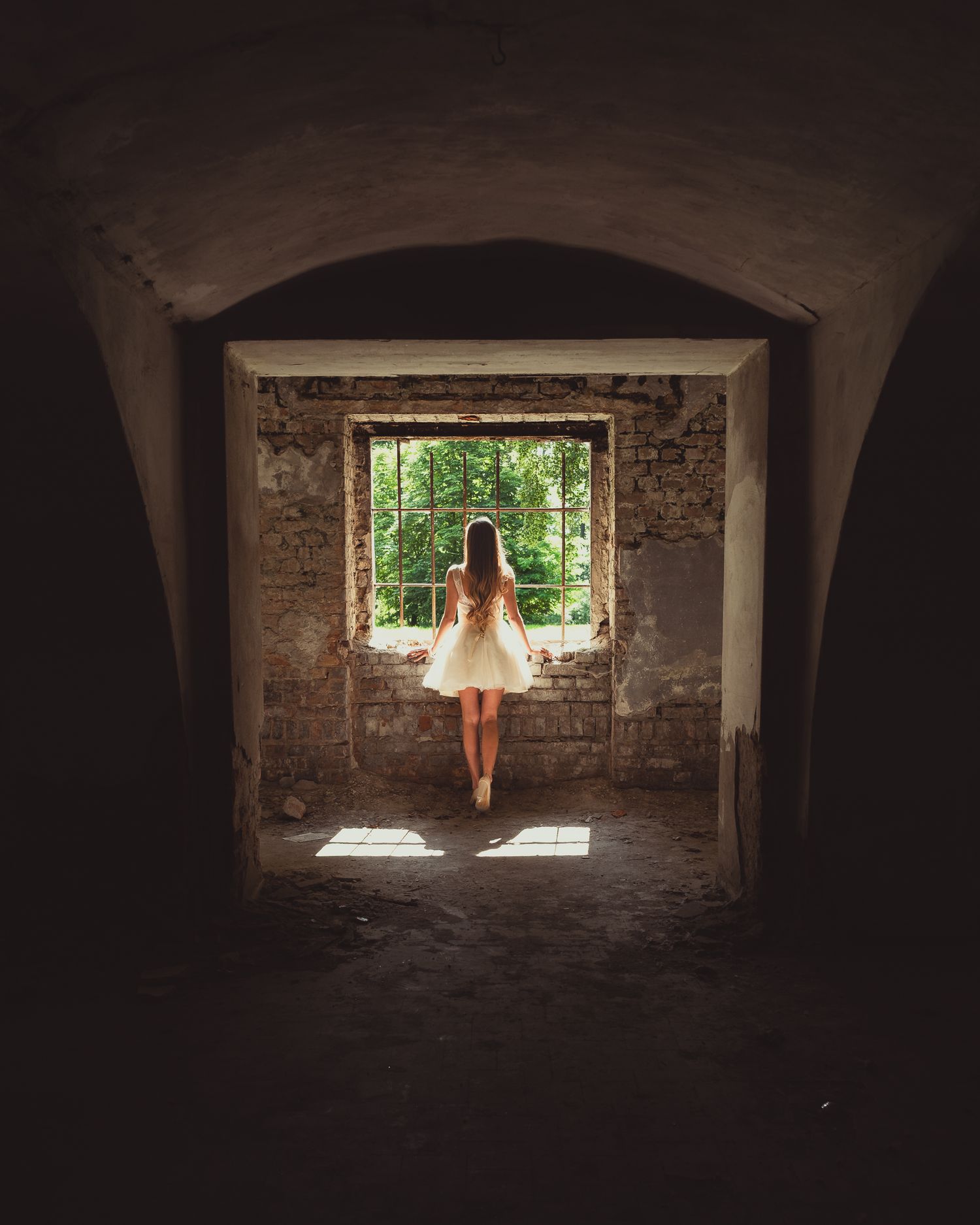 Editorial fashion: model in short tulle dress silhouette against barred window of abandoned brick vault, natural backlighting.