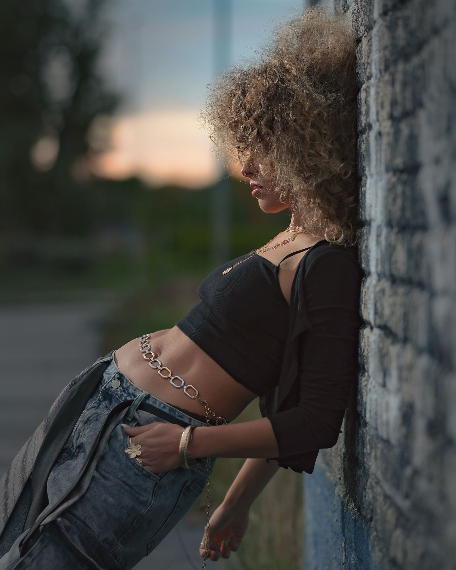Young woman with curly hair leaning against a brick wall at dusk, wearing a black crop top and jeans.