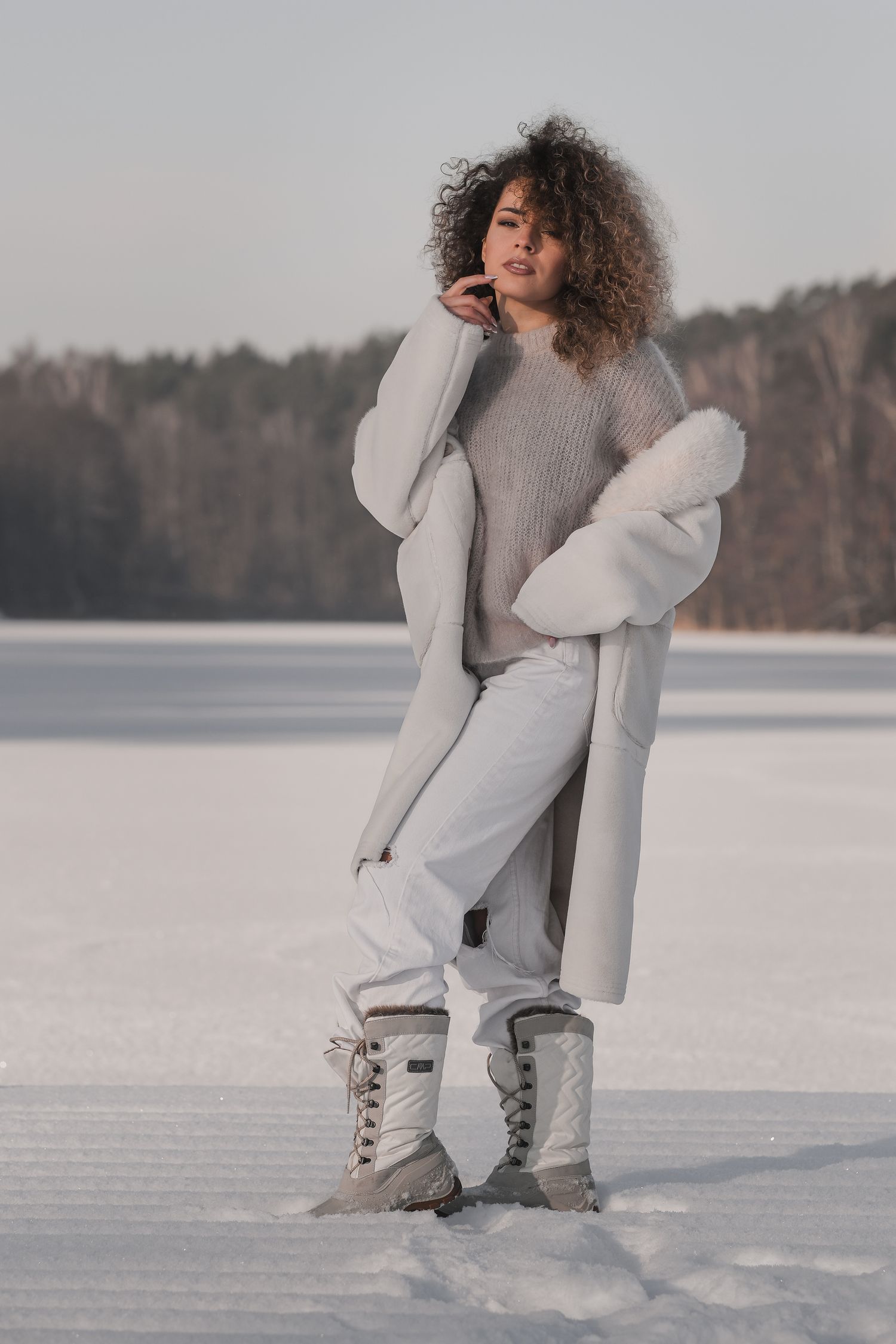 Full-body fashion editorial: model in white shearling coat, distressed jeans, and quilted snow boots, posing confidently on a snowy molo.