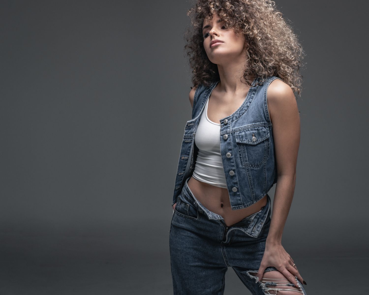 Woman with curly hair wearing a denim vest and jeans posing confidently against a gray background.