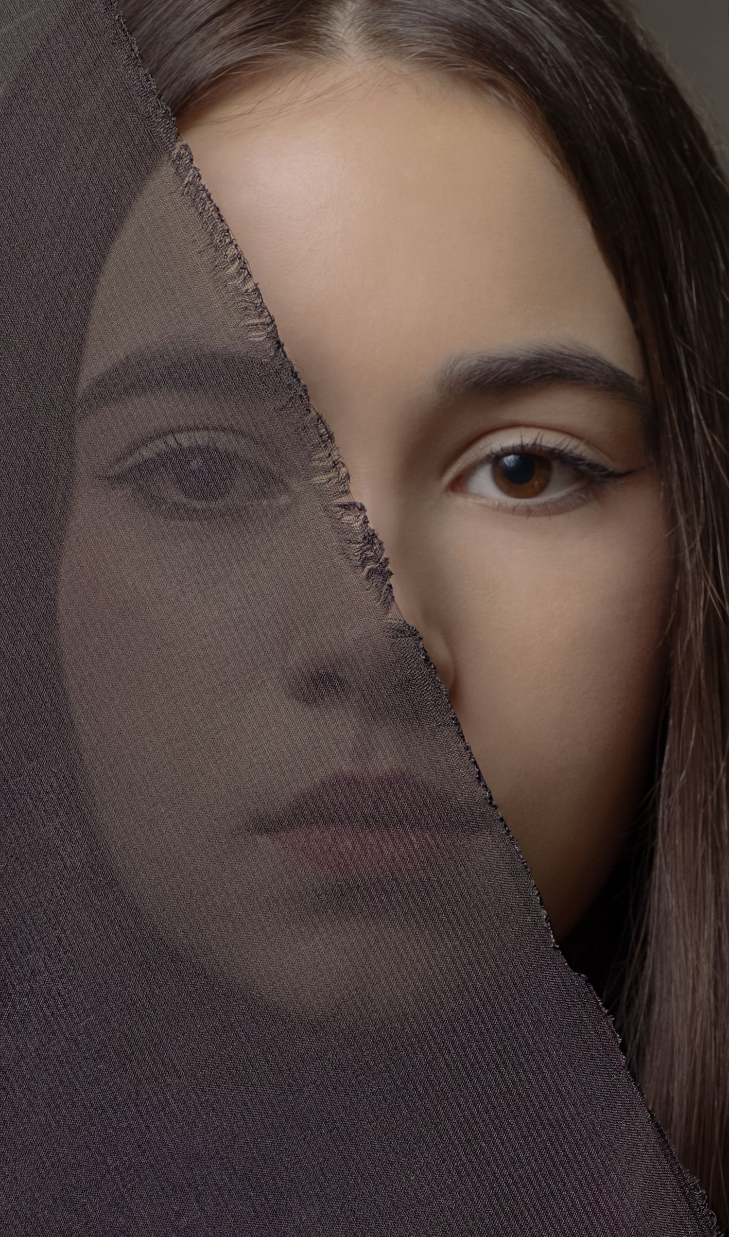 A young woman's face partially obscured by a dark fabric veil, creating a split artistic portrait effect.