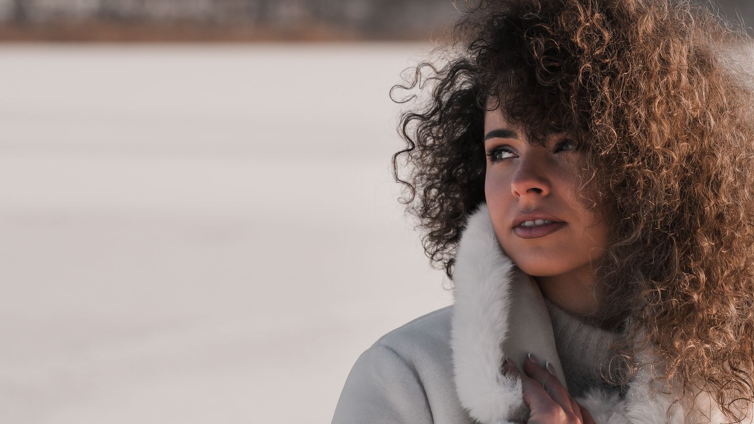 Global fashion photography: model profile shot with curly hair, touching white fur collar of a long coat, minimalist winter setting.