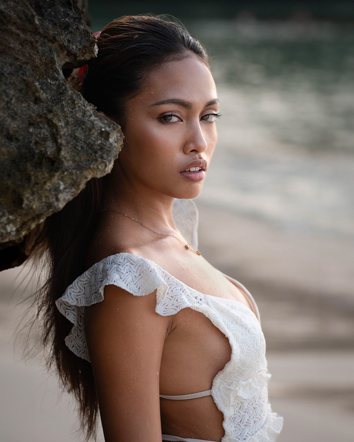 Professional beach editorial: close-up model in white lace top leans against coral rock, intense gaze, soft coastal lighting.