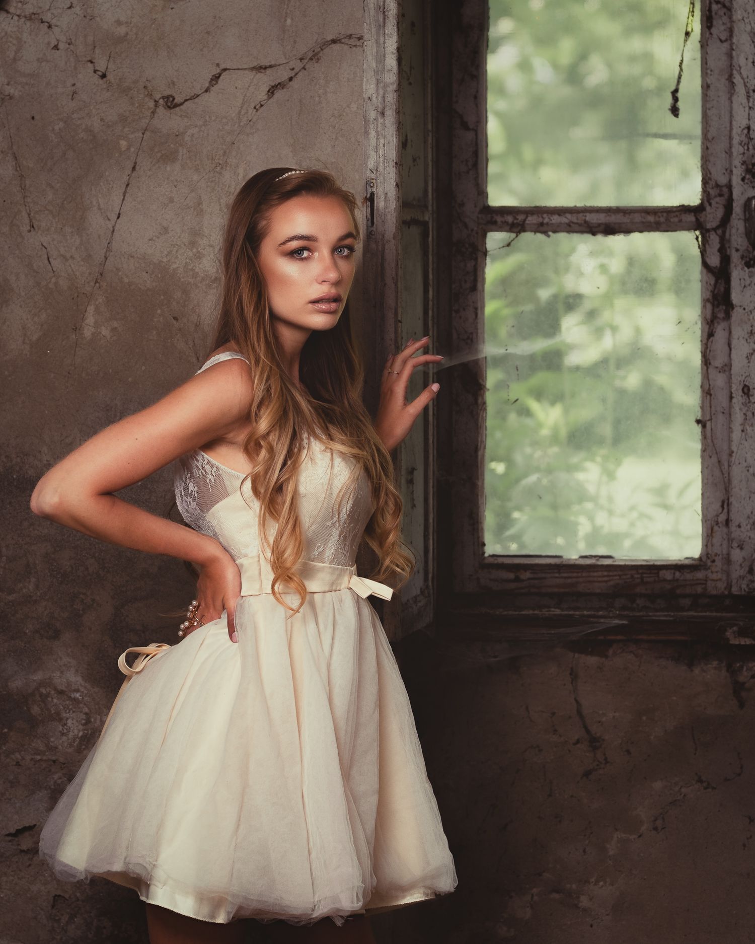 Editorial shot: model in short tulle dress by cobwebbed window, hand on hip, natural diffused light, rustic stone background texture.