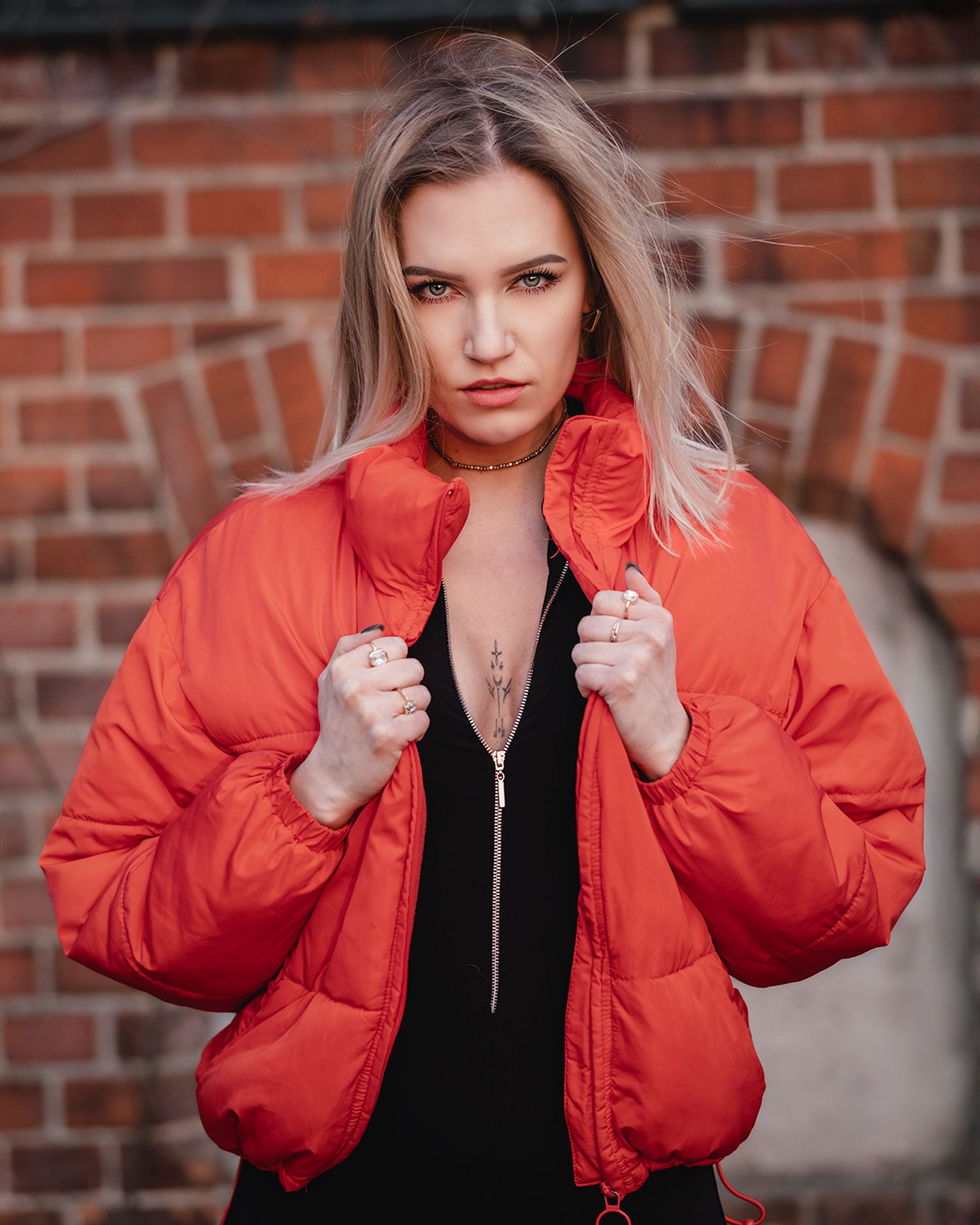 Blonde woman in red puffer jacket posing confidently against a brick wall in natural light.