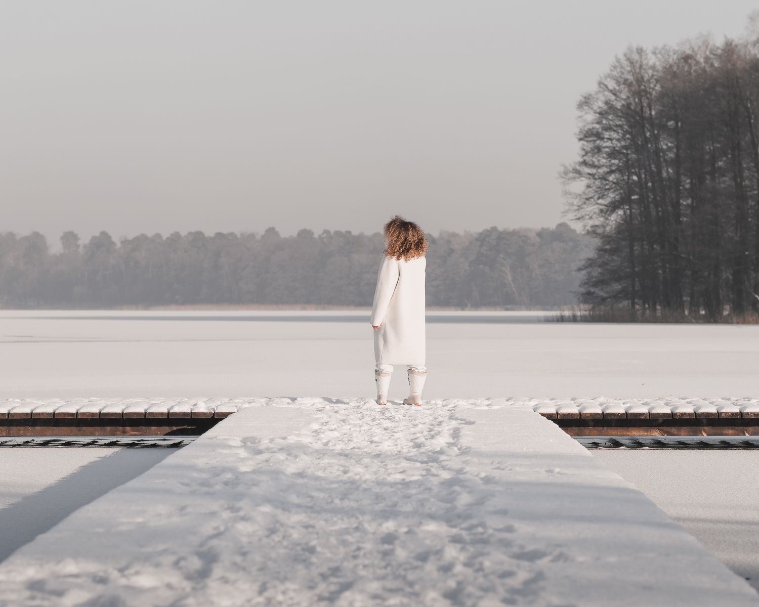 Editorial fashion: distant view of model in white coat walking on snow-covered wooden pier towards a frozen lake, scenic winter background.