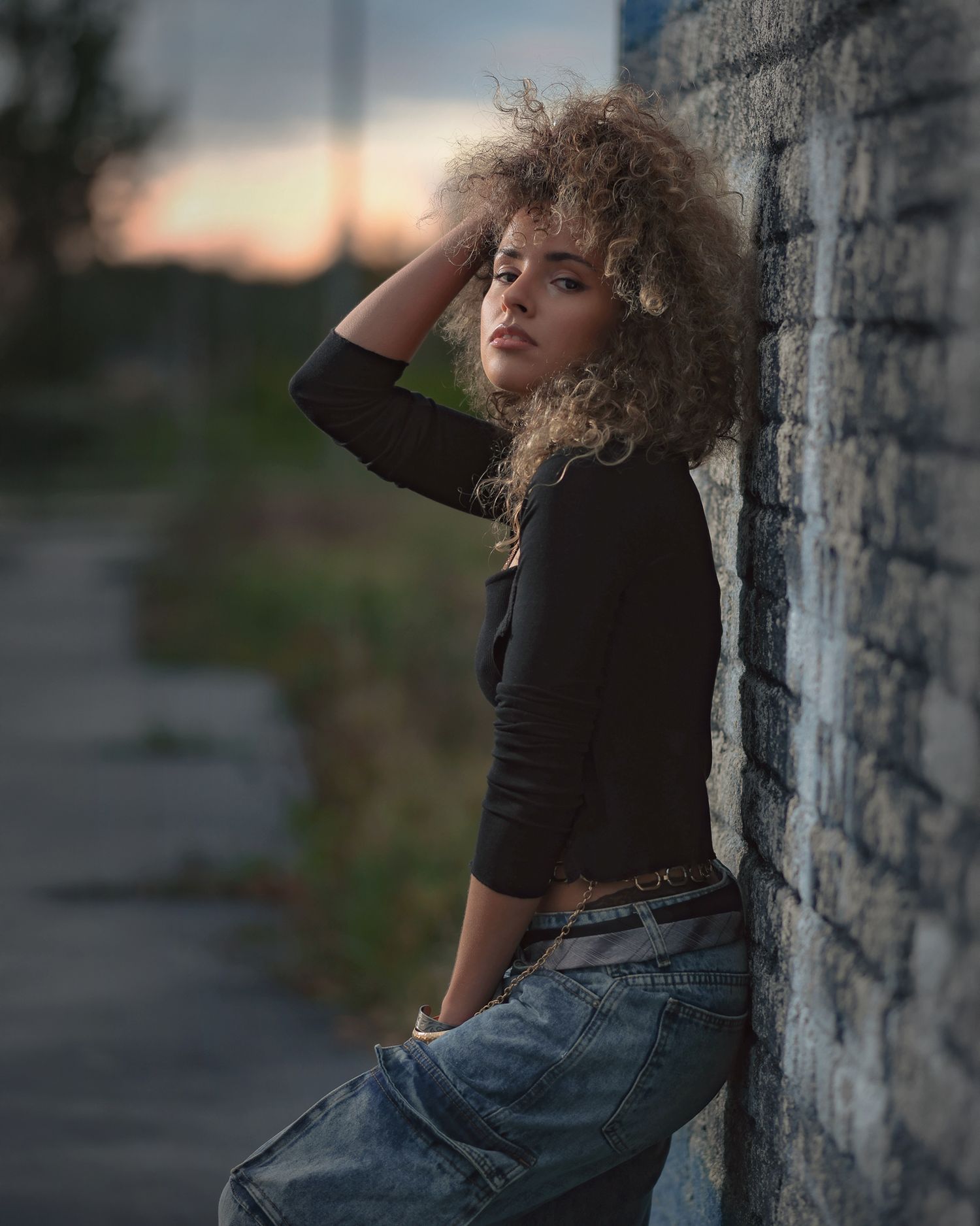 Young woman with curly hair leans against brick wall at dusk, wearing black top and jeans.