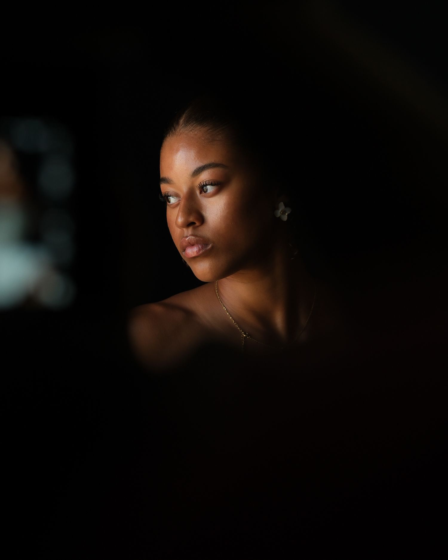 Young Black woman with a delicate necklace and earrings, dramatically lit against a dark background.