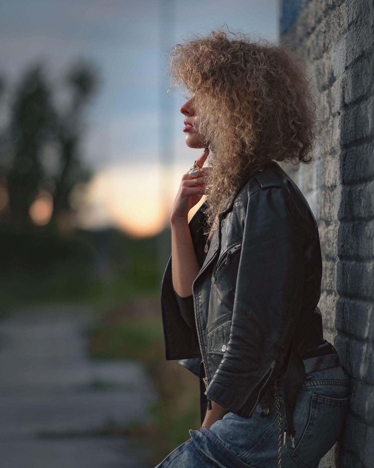 Urban fashion editorial: model with voluminous curly hair in a black leather jacket and jeans leaning on a rustic brick wall.