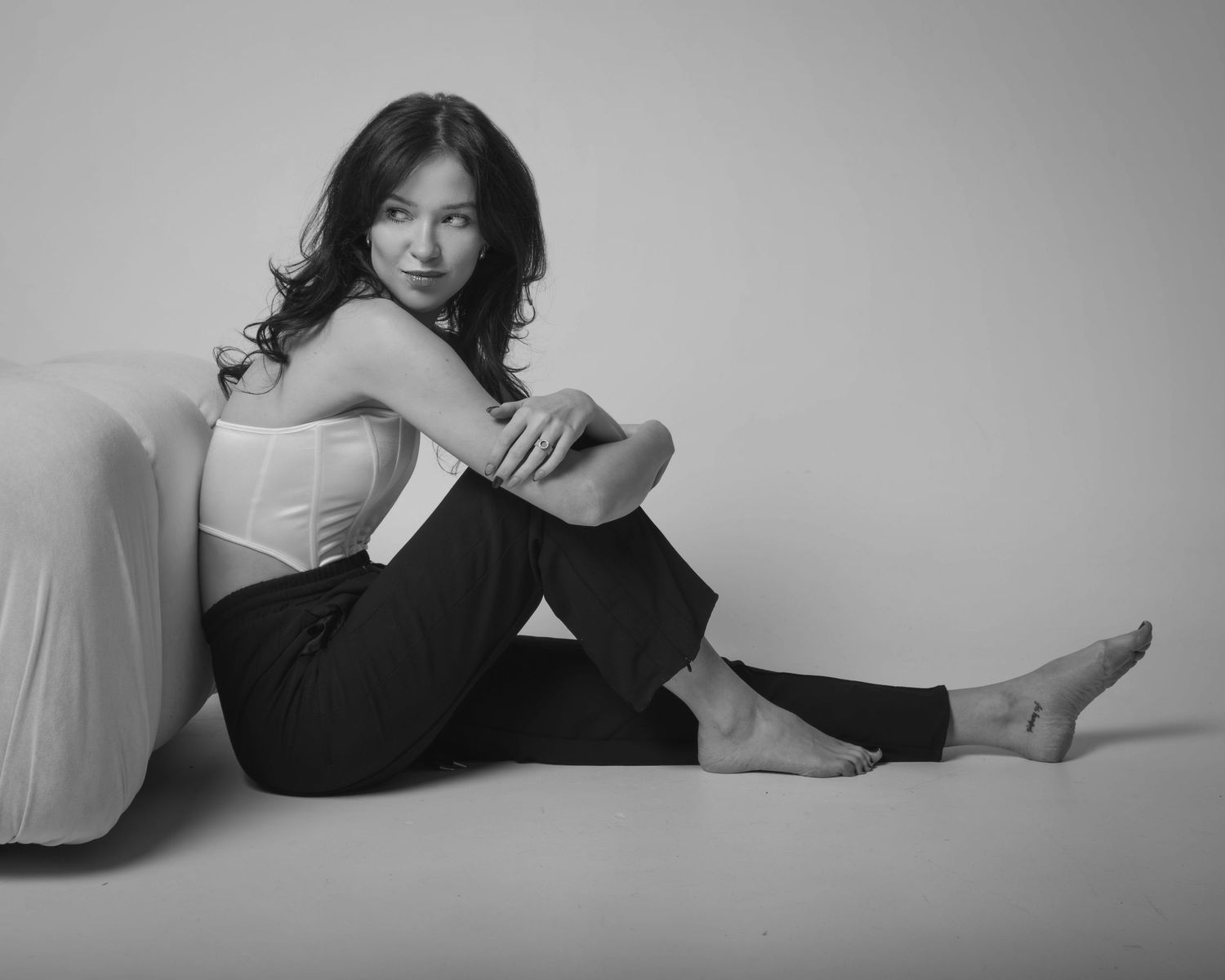 Full-body black and white photography: model barefoot, leaning on a pillow, clean studio background, international fashion portfolio.
