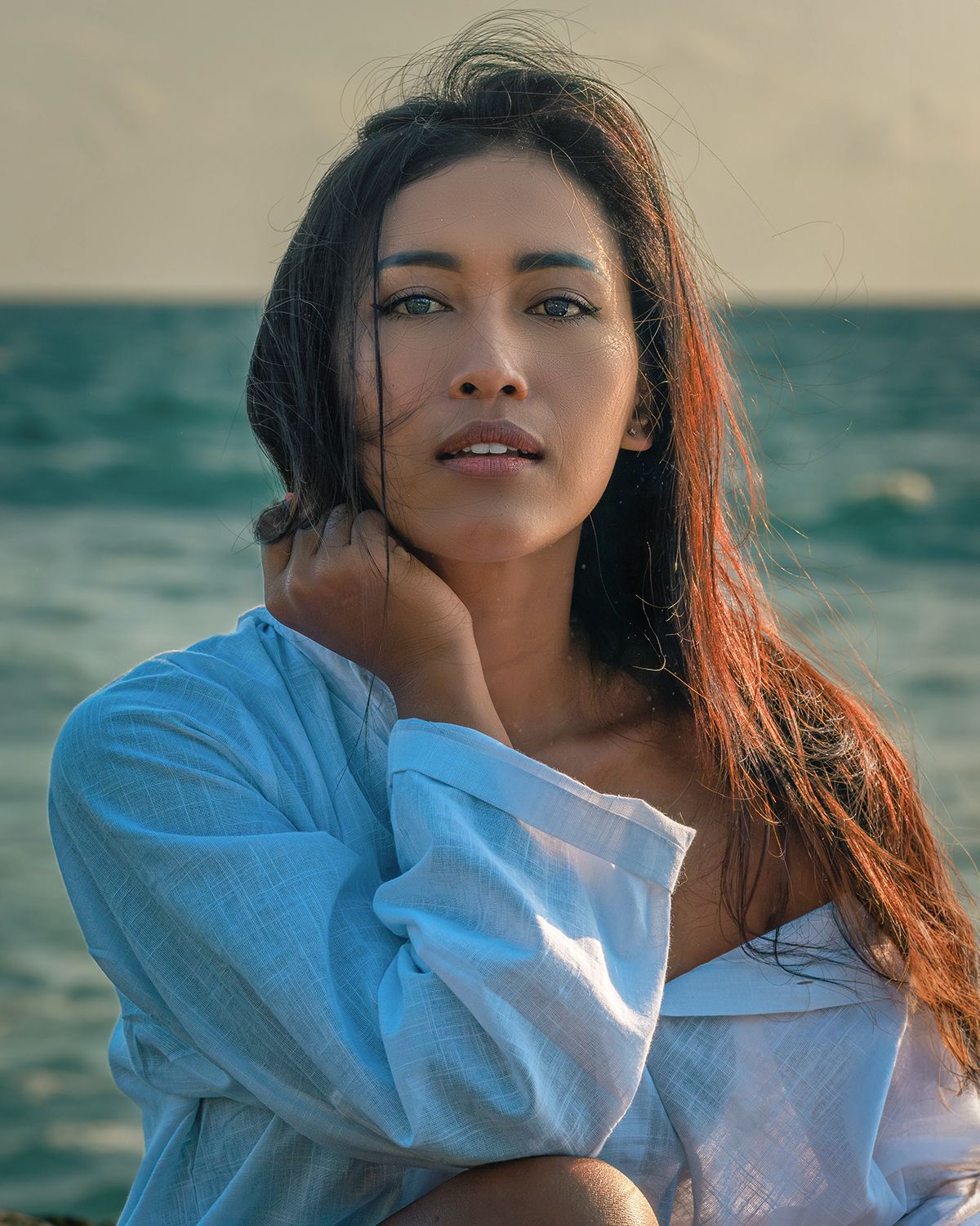 Young woman in white off-shoulder shirt poses by the ocean at sunset with long dark hair.