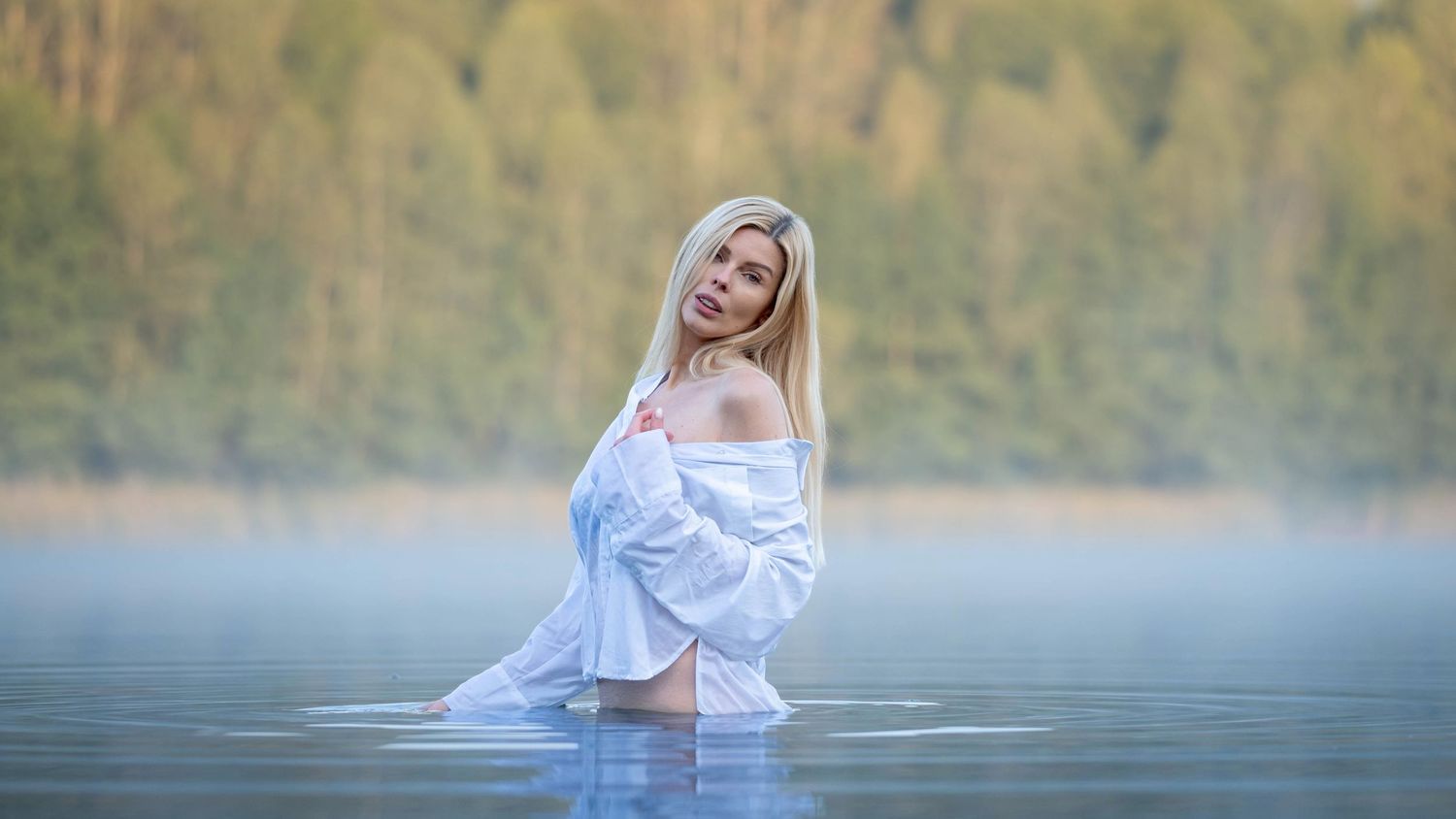Blonde woman in white off-shoulder shirt standing in misty lake water with forest background.