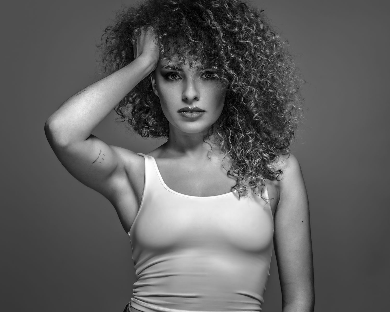 Professional black and white fashion shot: model with voluminous curly hair, posing with hand on head, subtle tattoo visible.