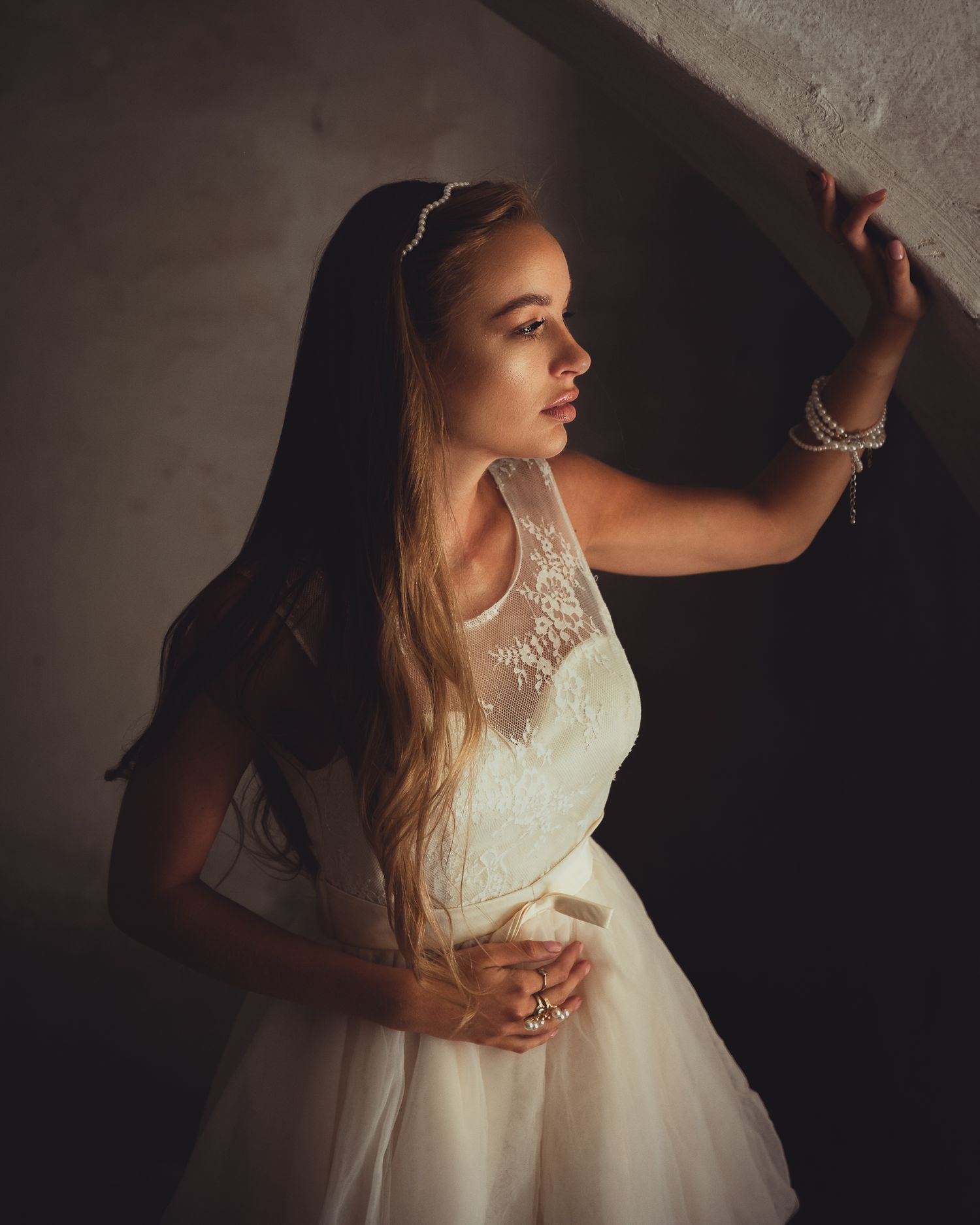 Cinematic fashion portrait: model in lace bodice tulle dress looking up, hand on rough wall, strong light and shadow, dramatic mood.
