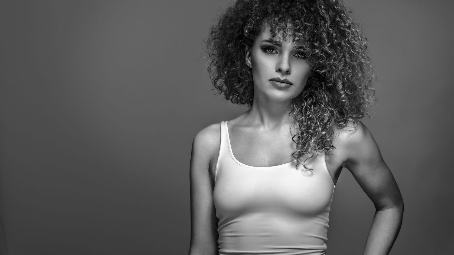 Black and white portrait of a young woman with curly hair wearing a white crop top, posing confidently.
