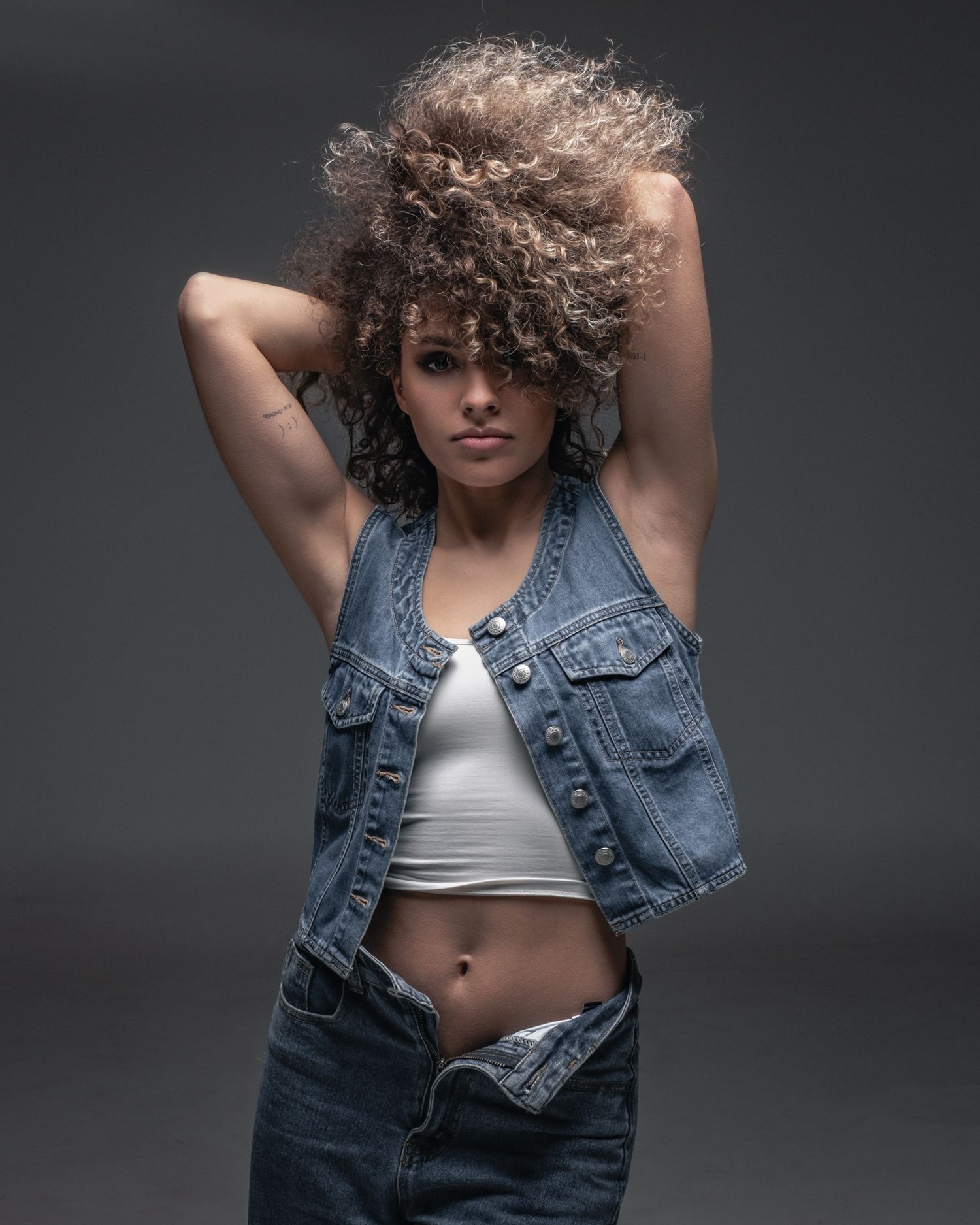 Mid-thigh fashion portrait: model with curly hair in a denim vest, dramatic studio shadows, minimalist aesthetic.