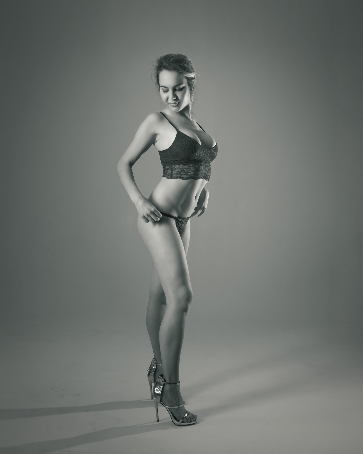 Black and white photo of a woman posing in lingerie and heels against a studio backdrop.